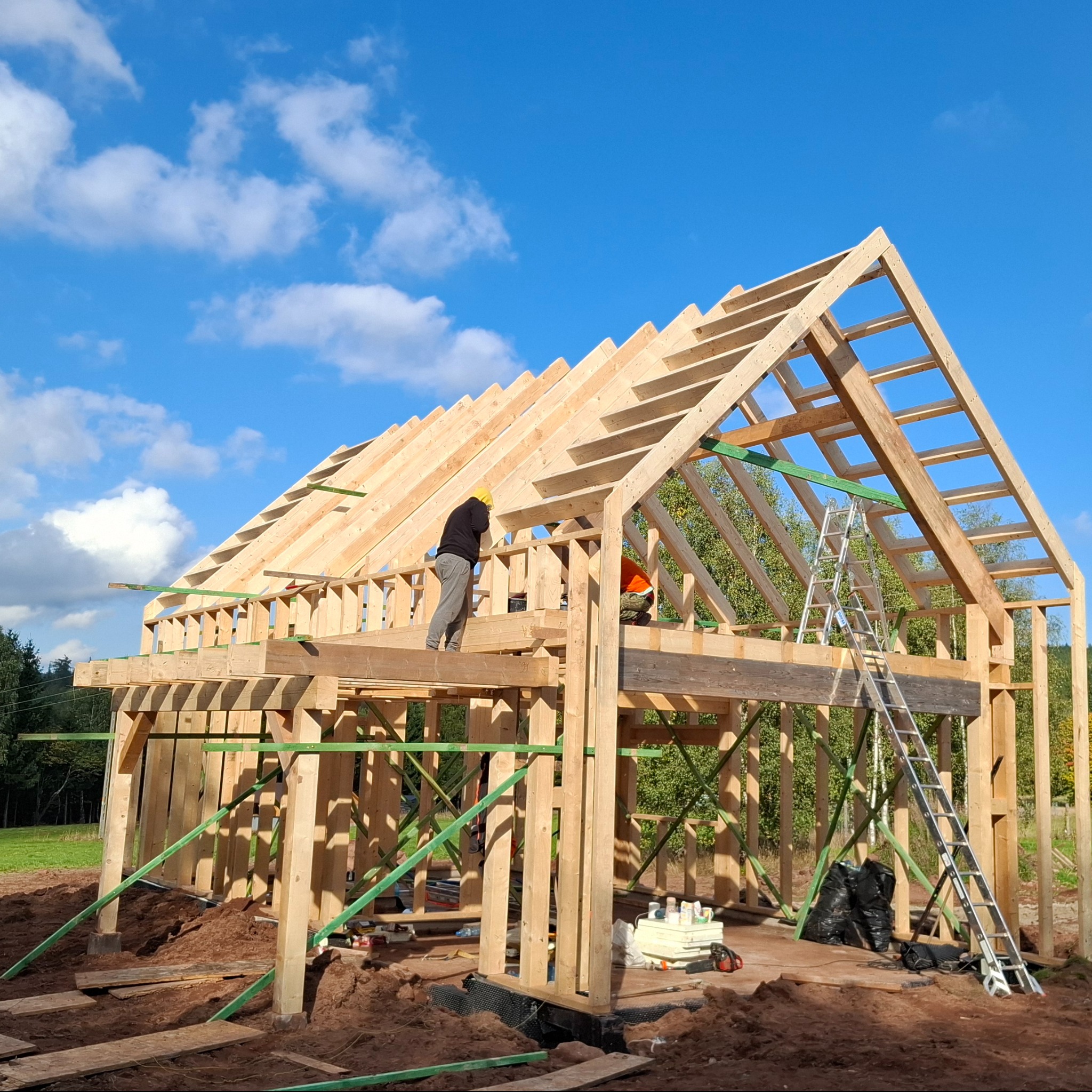 Wznoszenie drewnianej konstrukcji domu szkieletowego w słoneczny dzień. Widoczni pracownicy w kaskach, drabina oparta o konstrukcję, błękitne niebo z chmurami w tle.