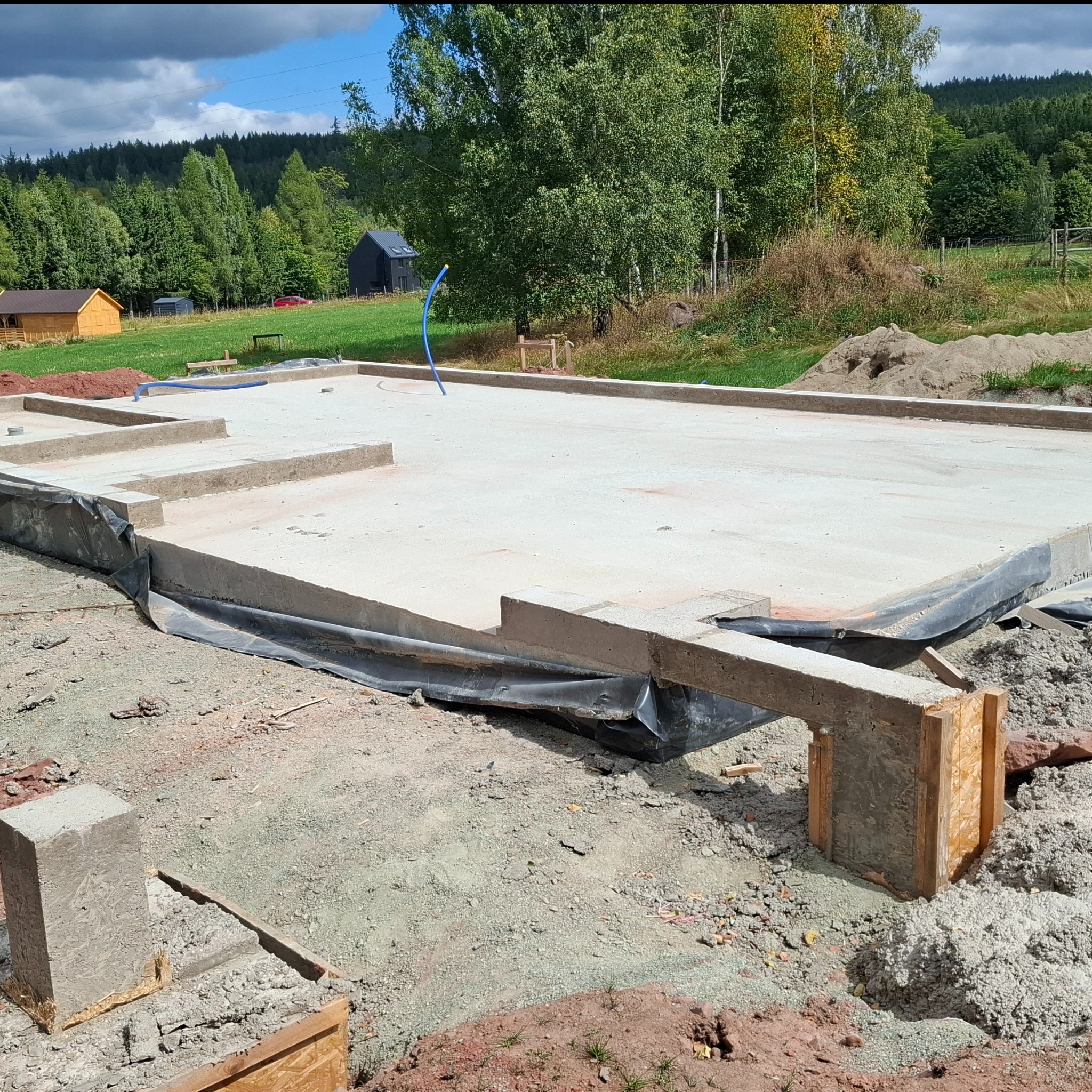 Wylana płyta fundamentowa z izolacją, widok z bliska na betonową konstrukcję i teren budowy. W tle zieleń drzew i zabudowania.