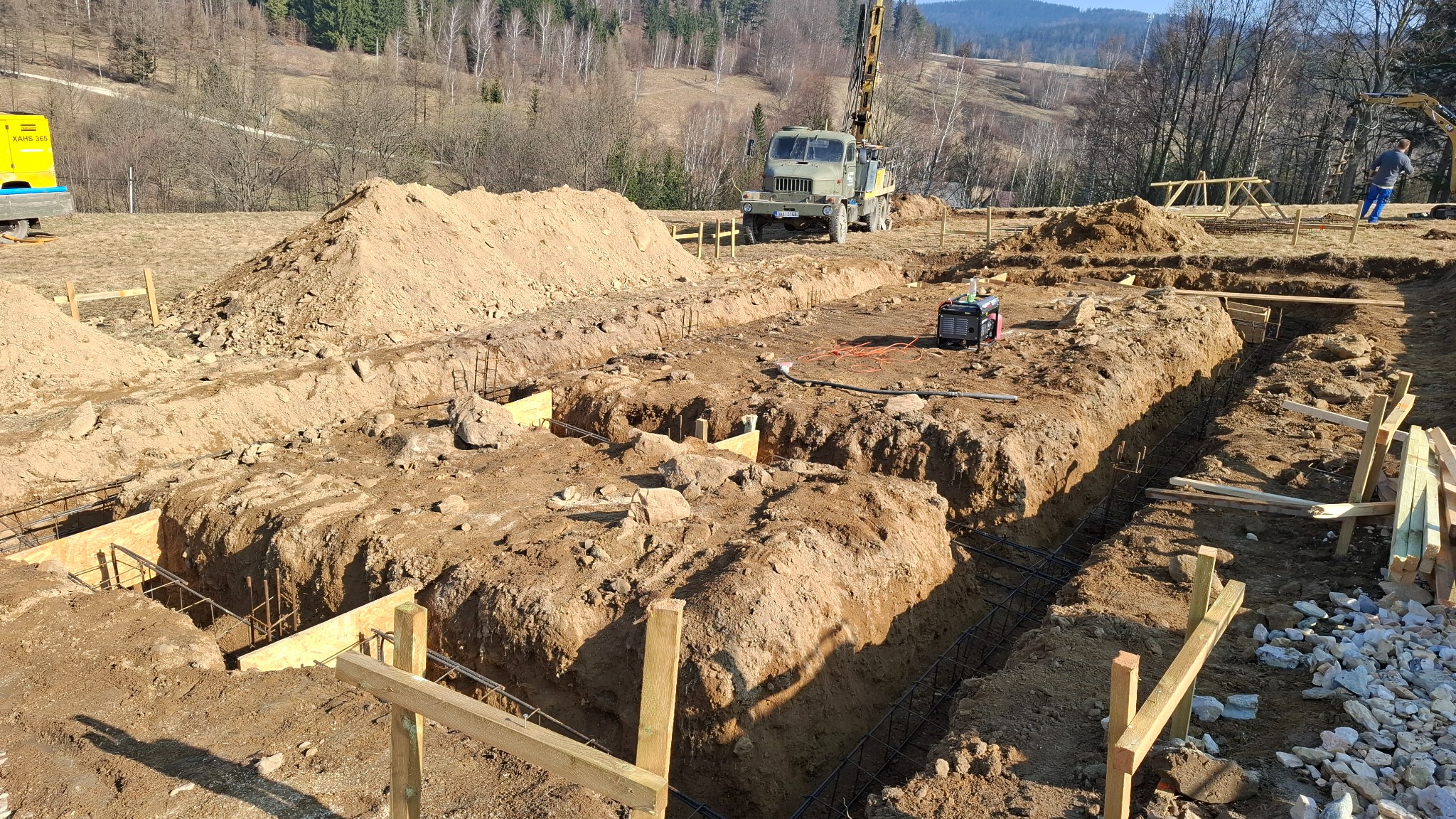 Wykopane rowy pod fundamenty z widocznym zbrojeniem, drewnianym szalunkiem i stertą ziemi w tle. Na drugim planie ciężarówka z wiertnicą i generator prądu.