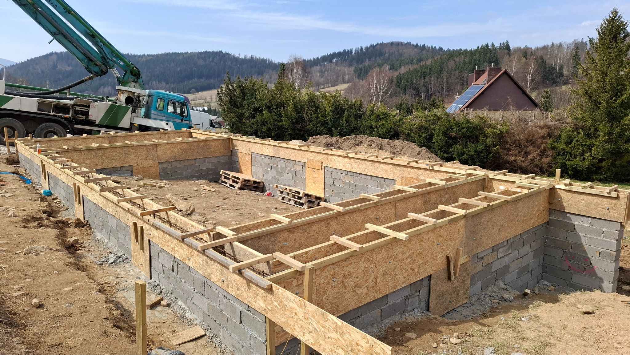 Wykopany fundament z szalunkiem i częściowo wymurowanymi ścianami z bloczków betonowych. W tle pompa do betonu i zalesione wzgórza.