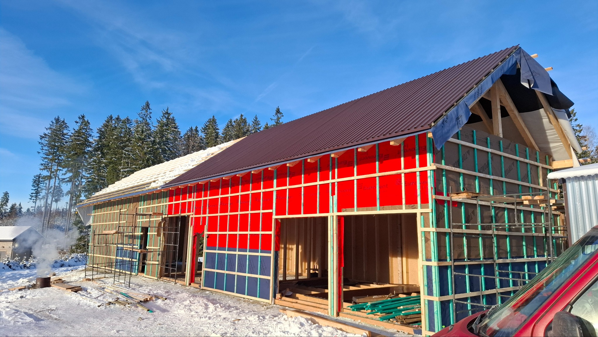 Wznoszenie domu szkieletowego w zimowej scenerii. Widoczne drewniane ramy, czerwone i niebieskie panele izolacyjne oraz częściowo ułożony brązowy dach. W tle ośnieżone drzewa.