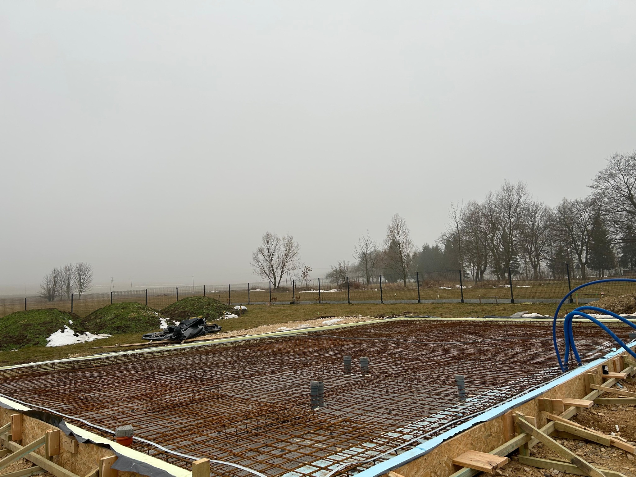 Zbrojenie fundamentu pod budowę domu, widoczne szalunki i wystające rury kanalizacyjne. W tle zieleń i mglista pogoda.