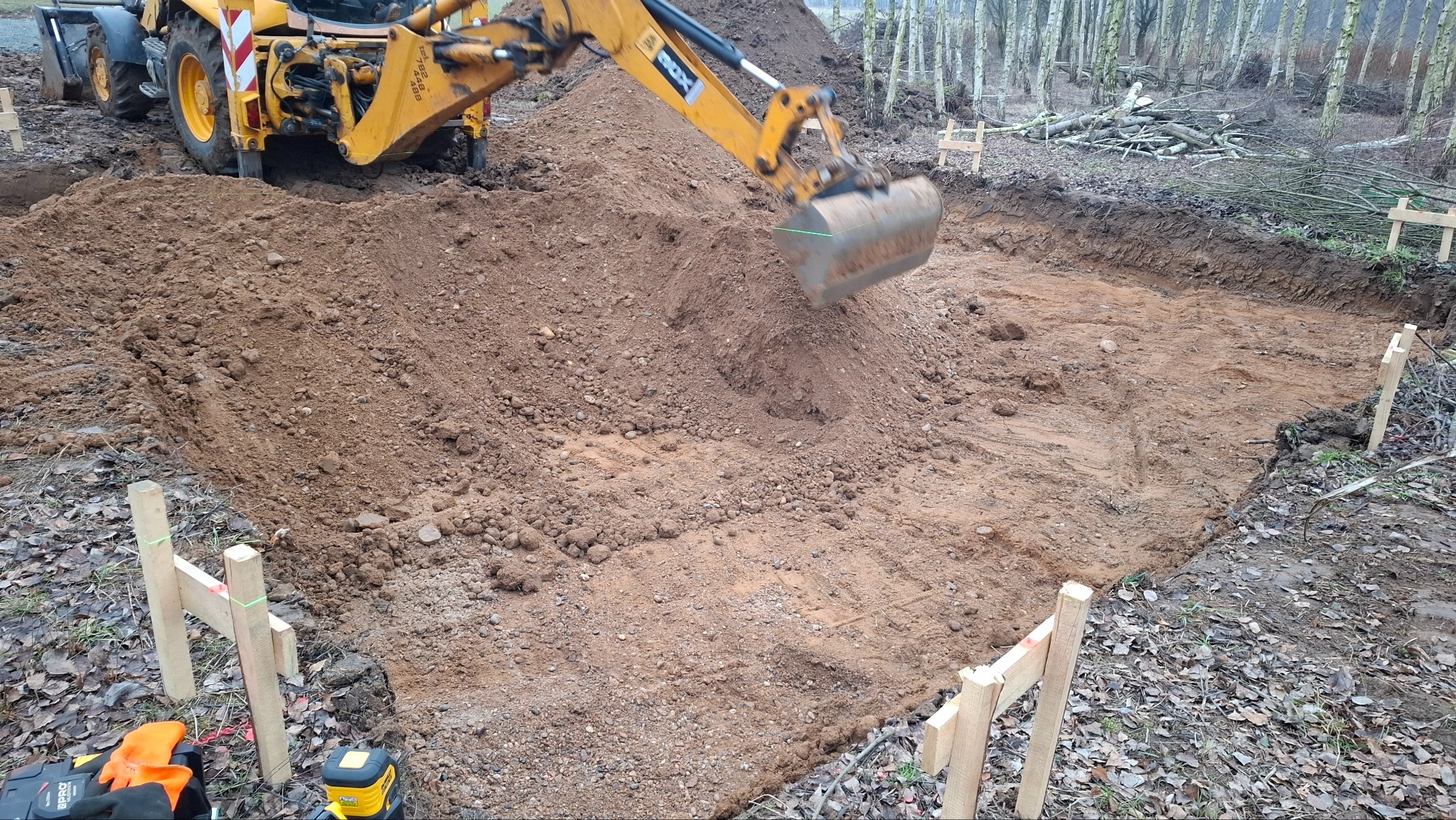 Żółta koparko-ładowarka wyrównuje teren pod budowę fundamentów, widoczne drewniane znaczniki i laserowy niwelator. Prace ziemne w toku, przygotowanie podłoża.