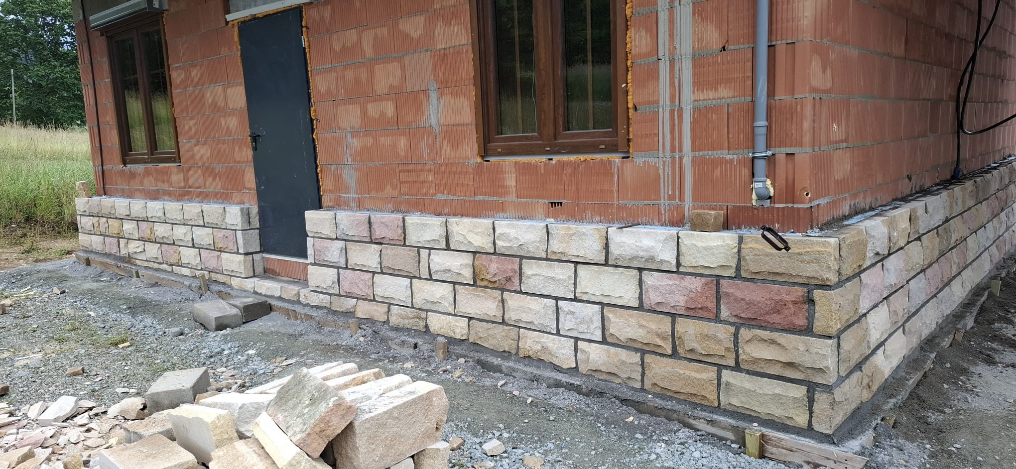 Wznoszenie muru z kamienia elewacyjnego na budynku z czerwonej cegły. Widoczne okno z brązową ramą i czarne drzwi. Częściowo wykonana elewacja z kamienia.