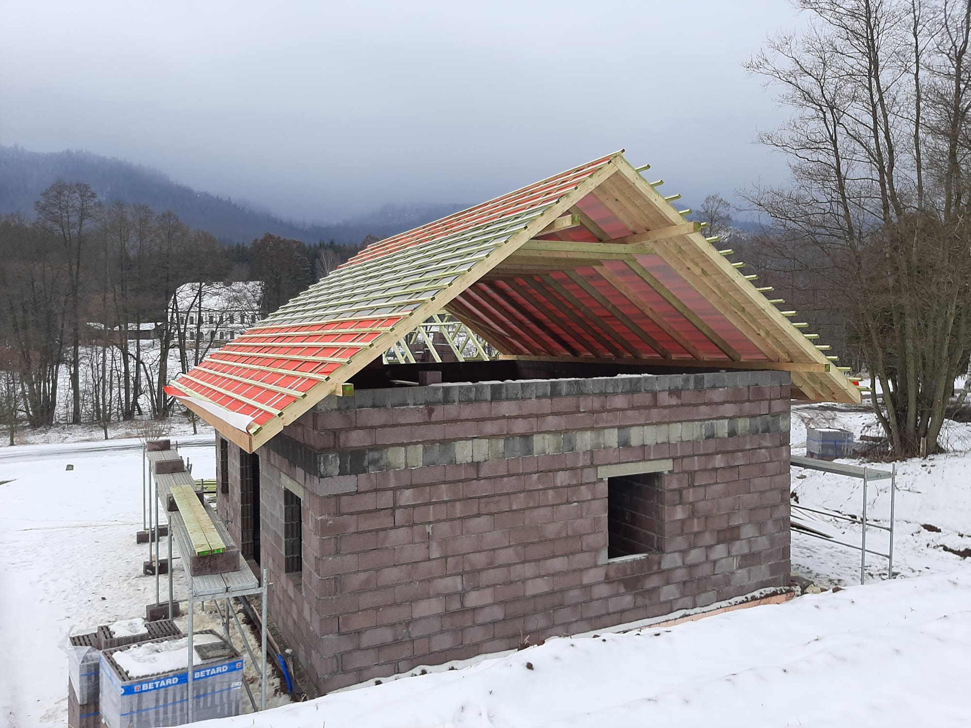 Budowa domu z bloczków: ściany wzniesione, konstrukcja dachu z drewnianych belek i czerwonej folii, rusztowania, zimowy krajobraz w tle. Etap surowy otwarty.