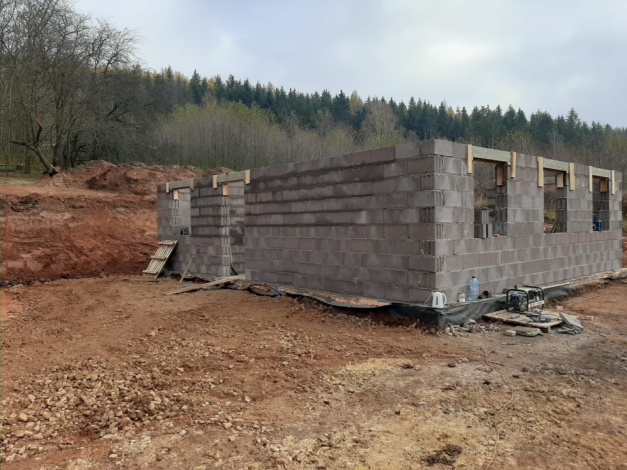 Wznoszenie ścian parteru z bloczków betonowych na fundamencie. Widoczne drewniane nadproża okienne i drzwiowe. W tle drzewa i ziemia.