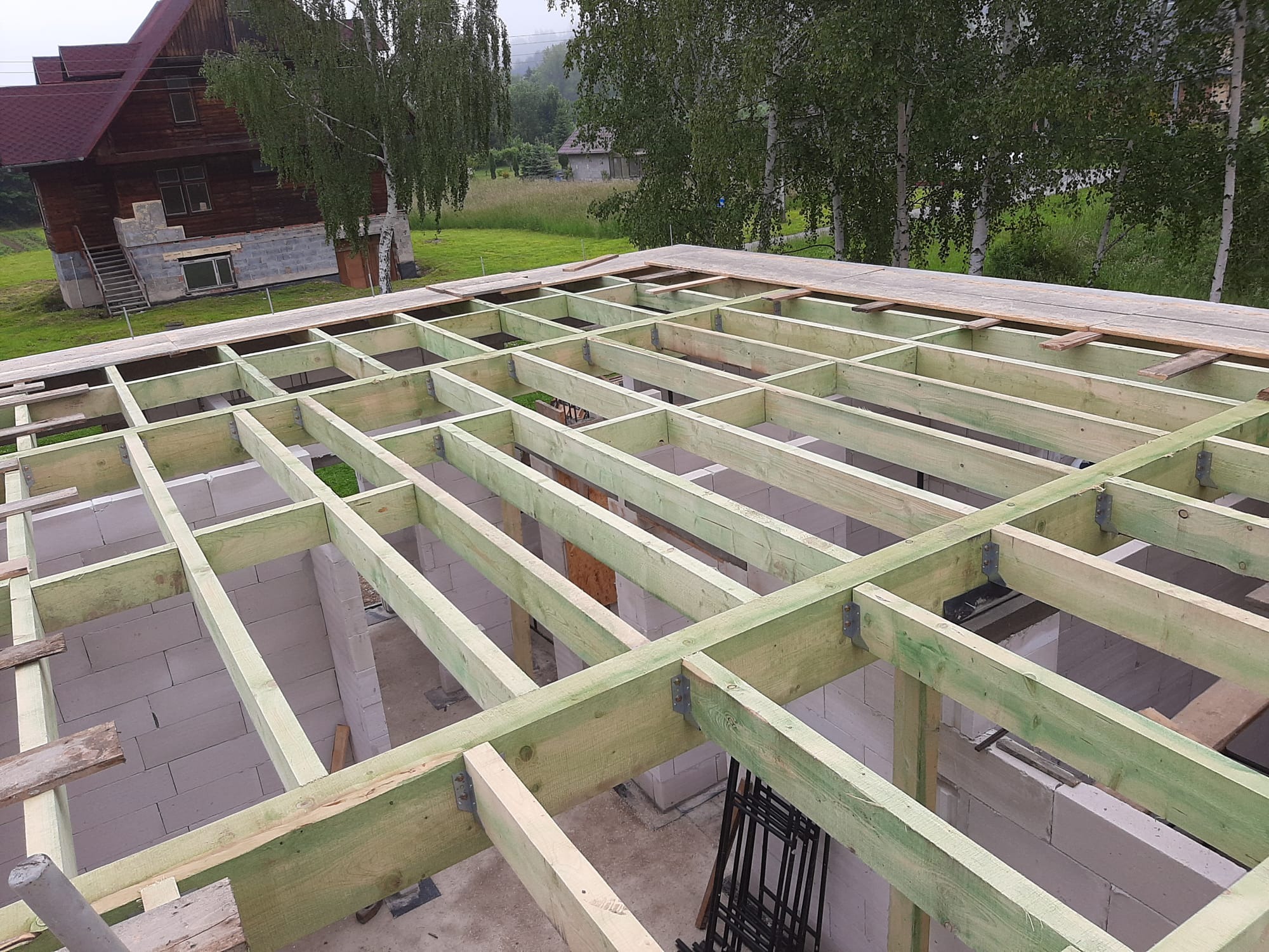 Konstrukcja drewniana dachu z jasnych belek, widok z góry na budynek w tle. Elementy połączone metalowymi wspornikami. Surowy stan, etap budowy.