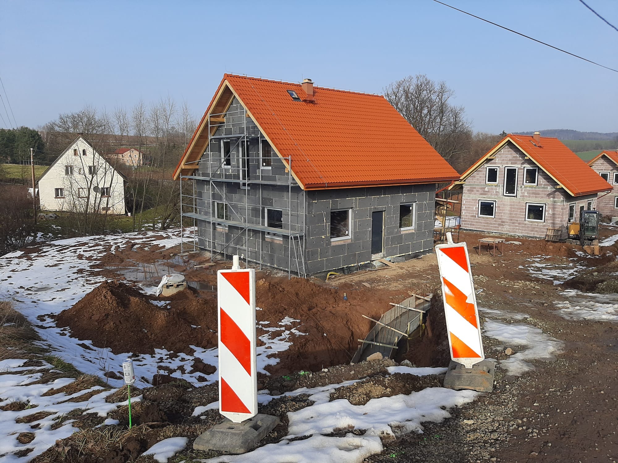Budowa domu z czerwonym dachem w Trzebieszowicach, widoczne rusztowania i fundamenty. W tle inne budynki w trakcie budowy, teren częściowo pokryty śniegiem.