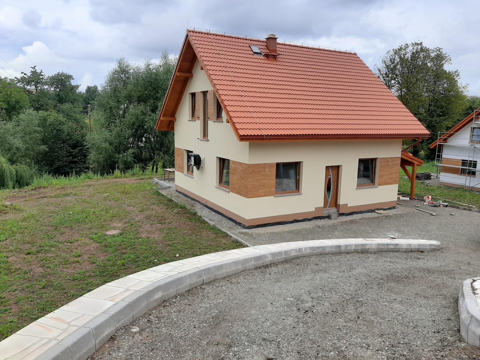 Ukończony dom z beżową elewacją i czerwoną dachówką, widok z zewnątrz, z elementami wykończenia imitującymi drewno. Widoczny fragment podjazdu z kostki.