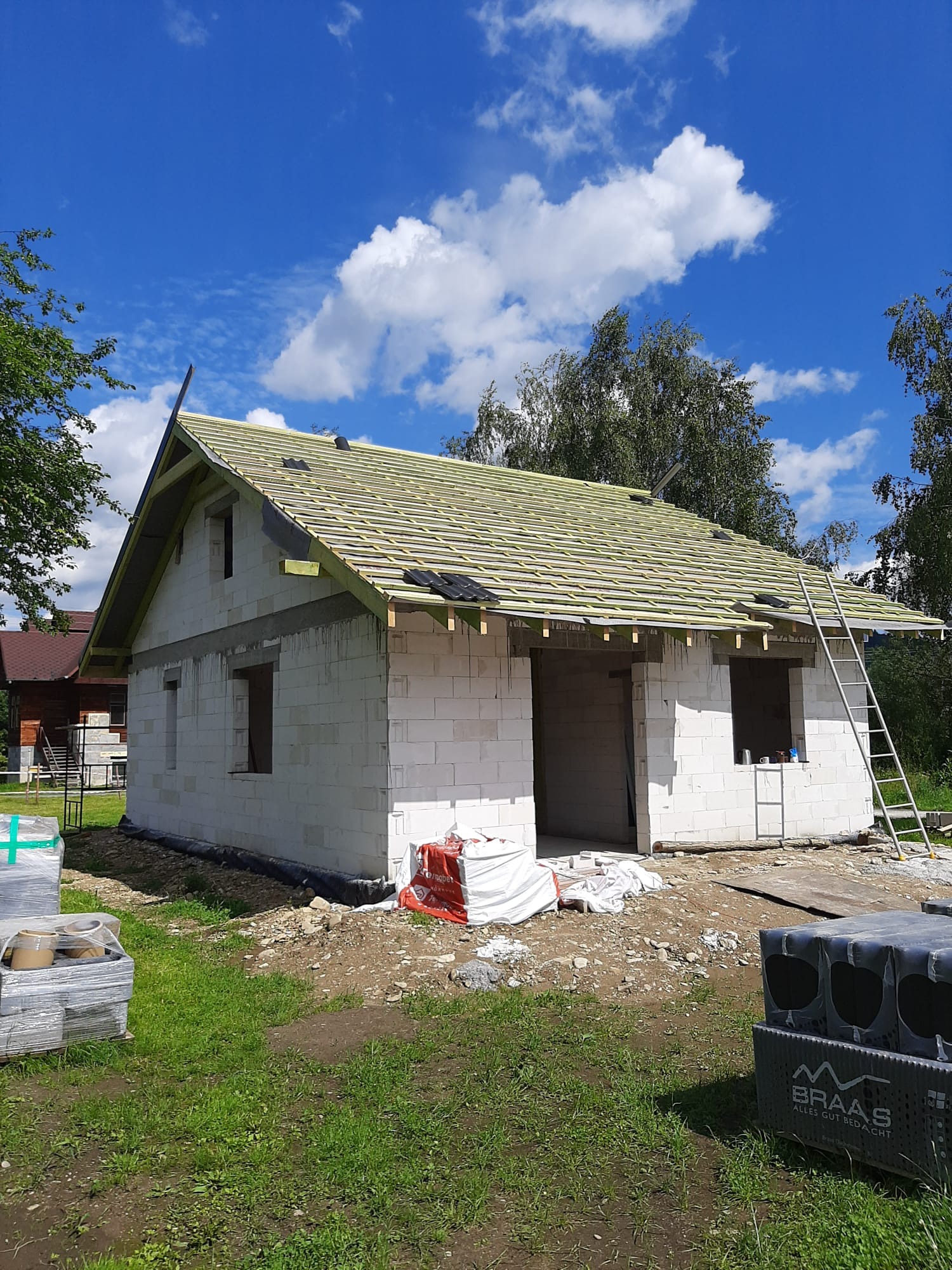 Budowa domu w trakcie: białe bloczki, drewniana konstrukcja dachu z łatami, drabina oparta o ścianę. Częściowo ułożone dachówki. Materiały budowlane na pierwszym planie.