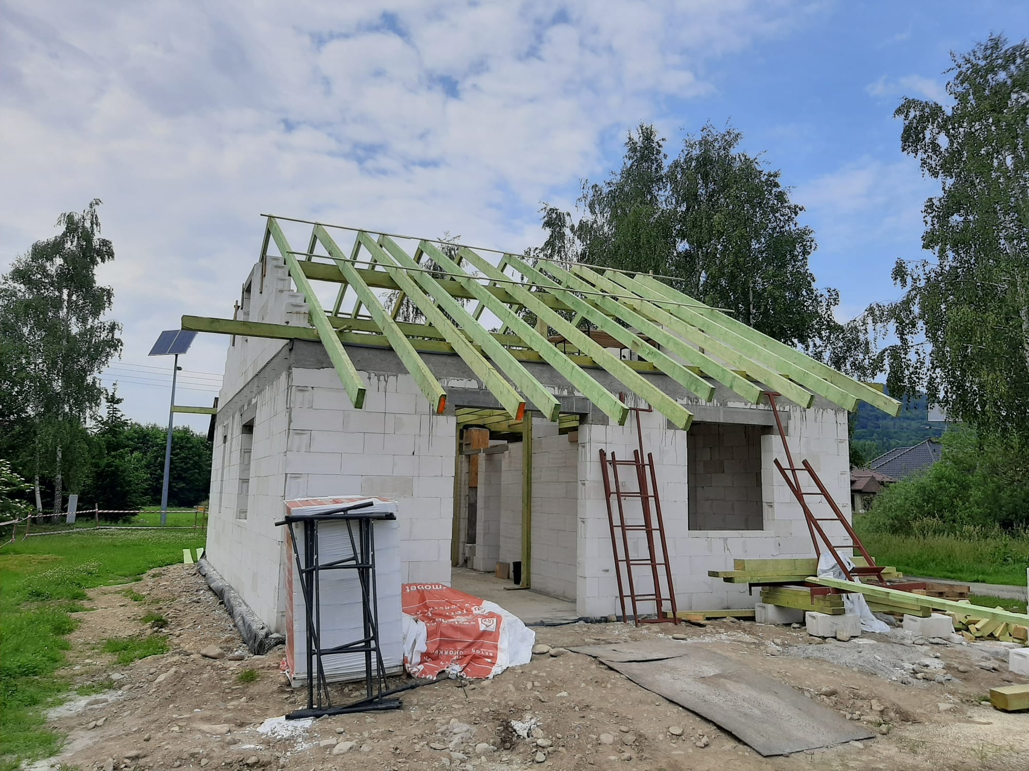 Budowa domu z białych bloczków: ściany wzniesione, zielone krokwie dachu, drabiny i materiały budowlane na placu. Widok na budynek w trakcie realizacji.