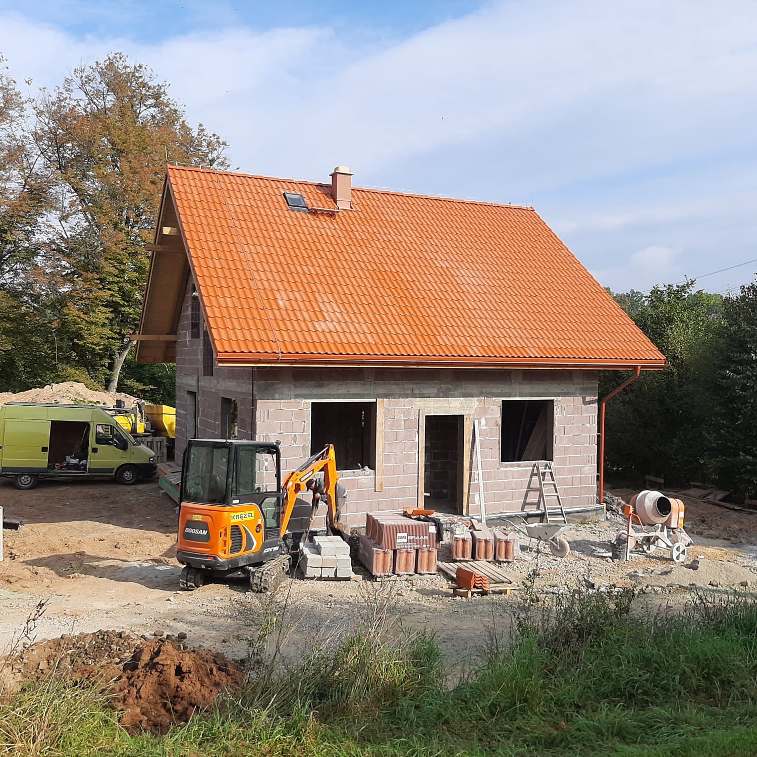 Budowa domu w trakcie: murowane ściany z pustaków, pomarańczowy dach z dachówki, mini koparka Doosan i betoniarka na placu budowy, częściowo widoczny żółty bus.