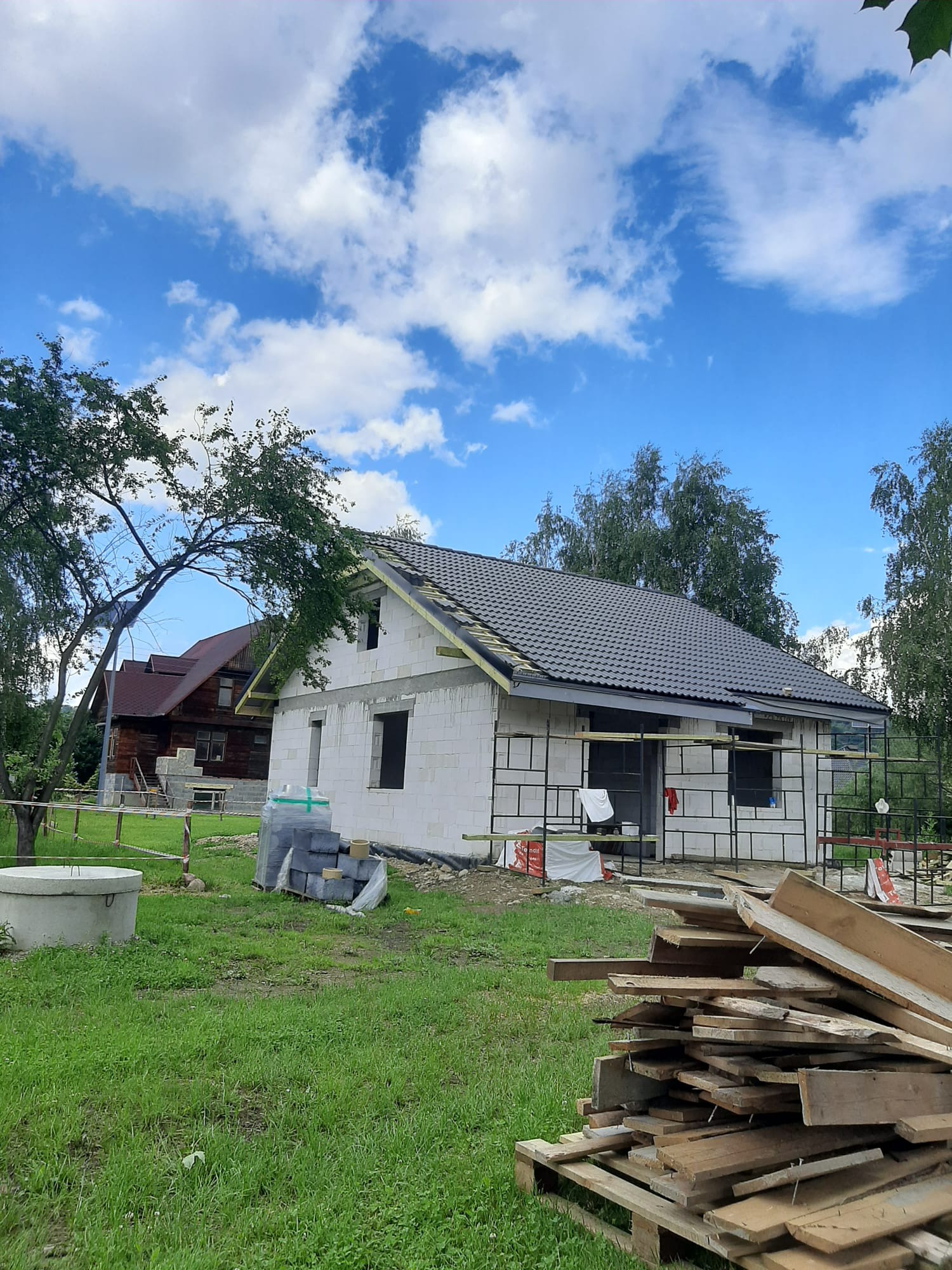 Budowa domu jednorodzinnego w trakcie realizacji: białe ściany z bloczków, szara dachówka, widoczne rusztowania i materiały budowlane na zielonej działce. Błękitne niebo.