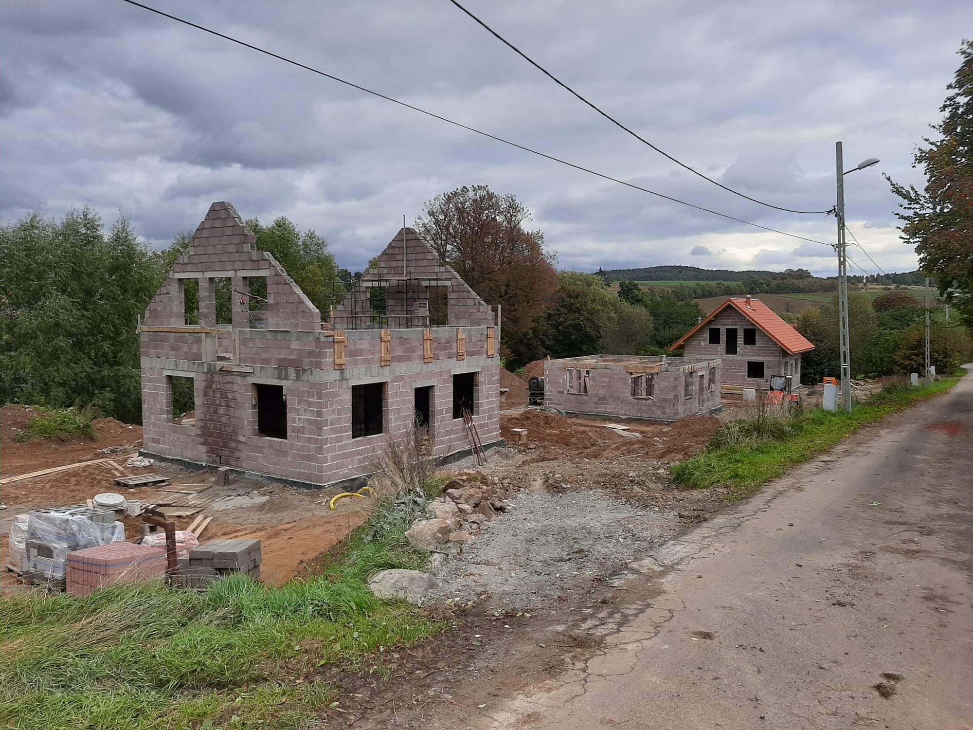 Budowa dwóch domów z betonu komórkowego w stanie surowym otwartym, widok z boku drogi, pochmurne niebo w tle. Częściowo ułożona dachówka na jednym z budynków.