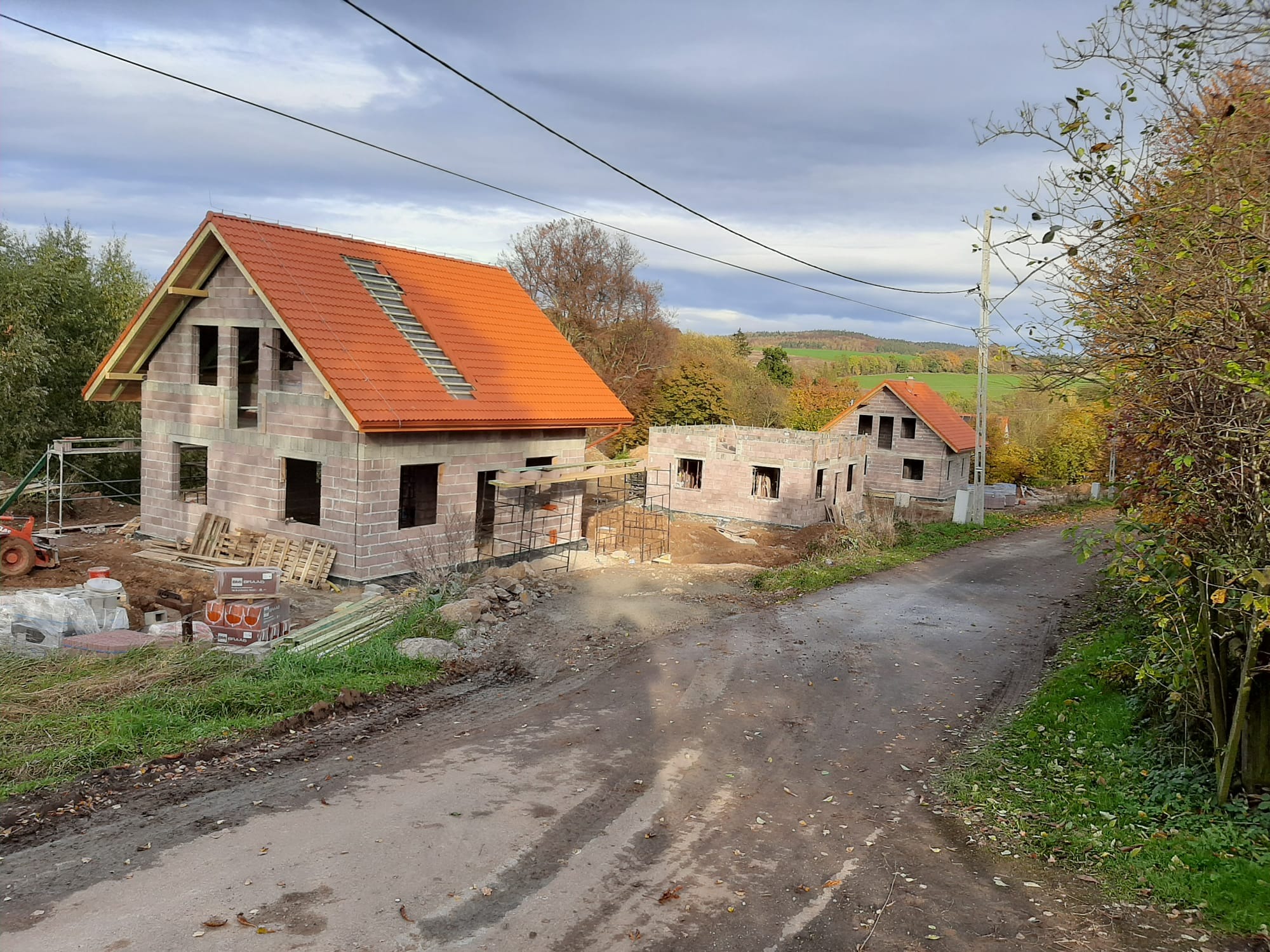 Dwa domy w stanie surowym otwartym z czerwoną dachówką, widoczne materiały budowlane i krajobraz w tle. Jeden dom z częściowo ukończonym dachem.