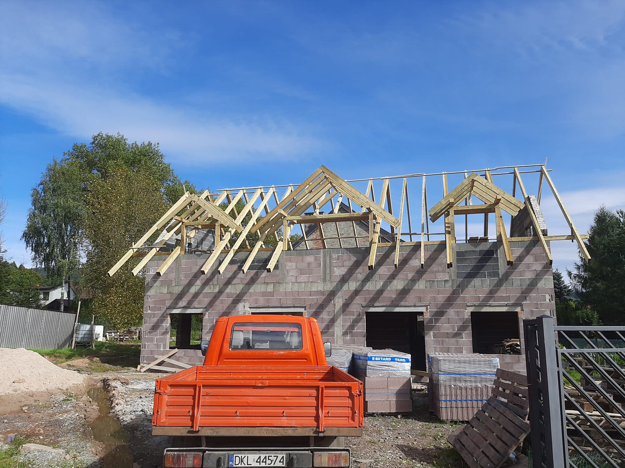 Budowa domu z bloczków: ściany z otworami okiennymi, więźba dachowa w trakcie montażu. Na pierwszym planie pomarańczowa ciężarówka i materiały budowlane.