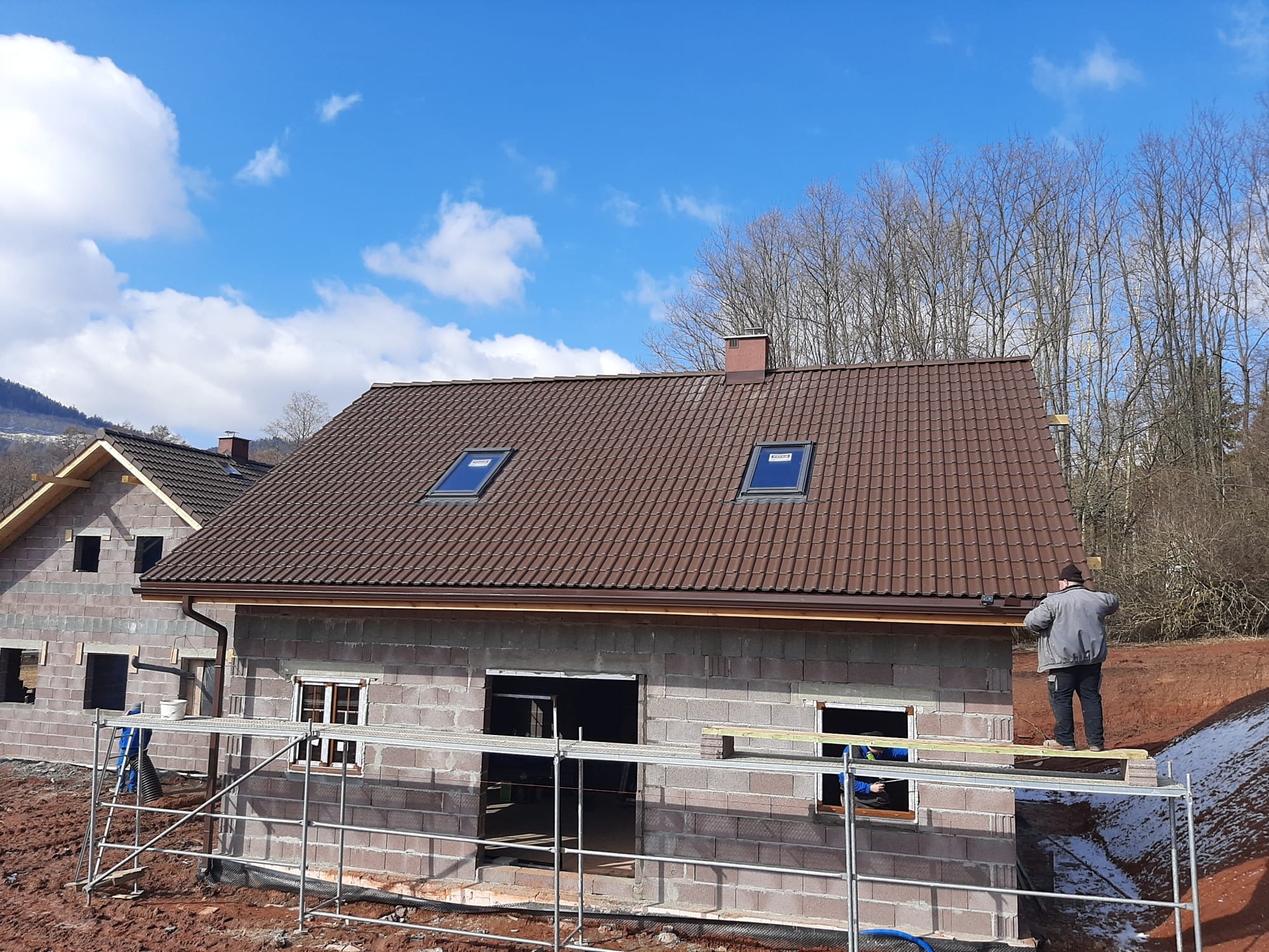 Budowa domu z bloczków: surowy stan z brązowym dachem i oknami dachowymi. Na rusztowaniu stoi pracownik. W tle drugi budynek i drzewa.