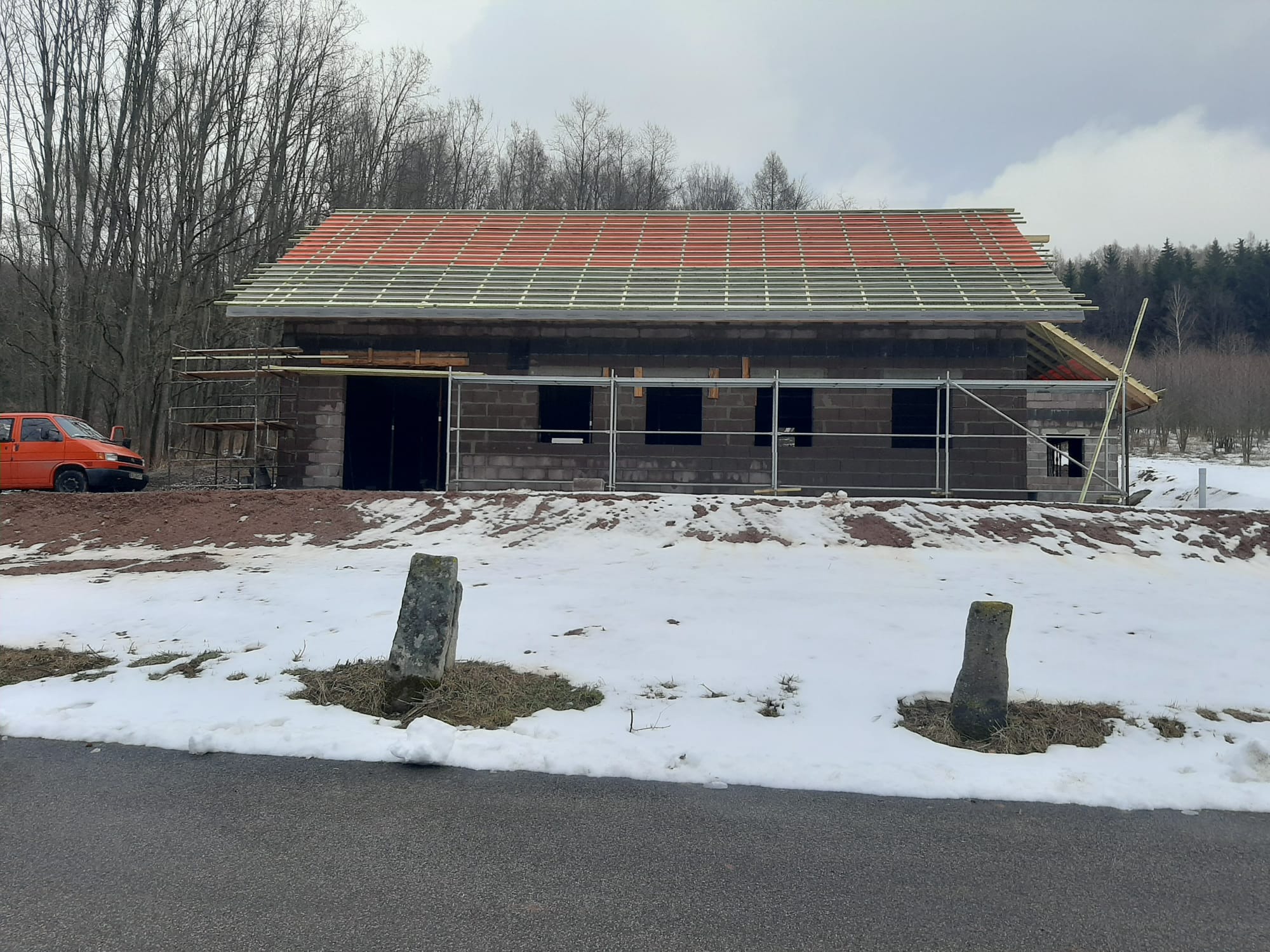 Budowa domu z pustaków w zimowym krajobrazie. Częściowo położony dach z czerwoną dachówką. Widoczne rusztowania i pomarańczowy samochód w tle.