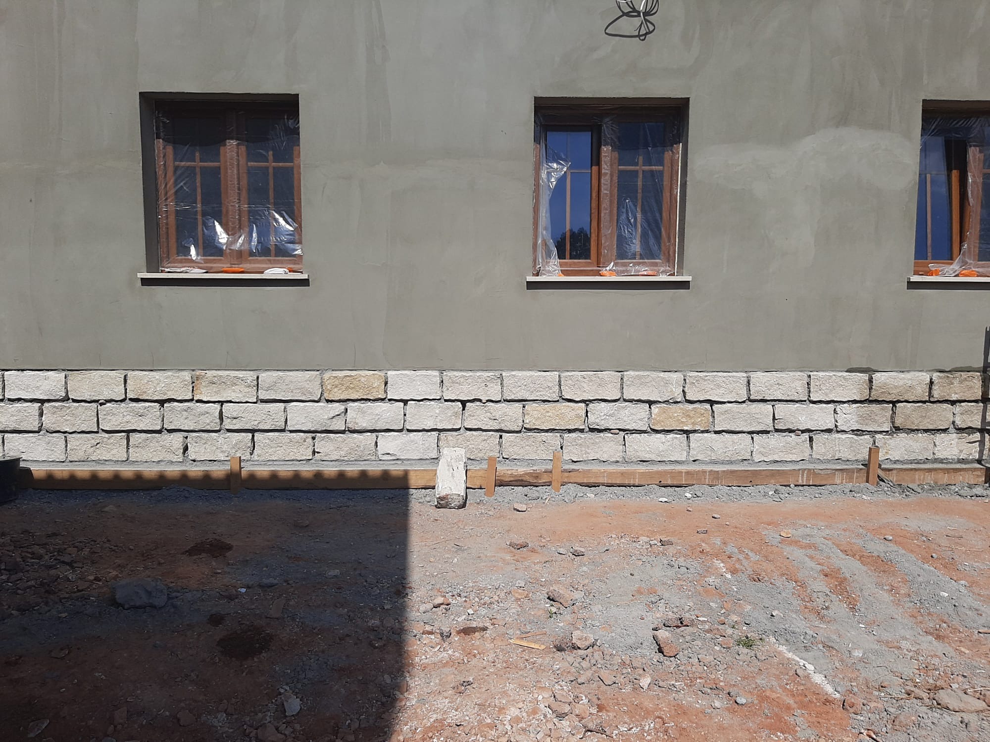 Ściana budynku z oknami zabezpieczonymi folią i fragmentem elewacji z kamienia. Widoczne drewniane elementy szalunku fundamentu. Prace w toku.