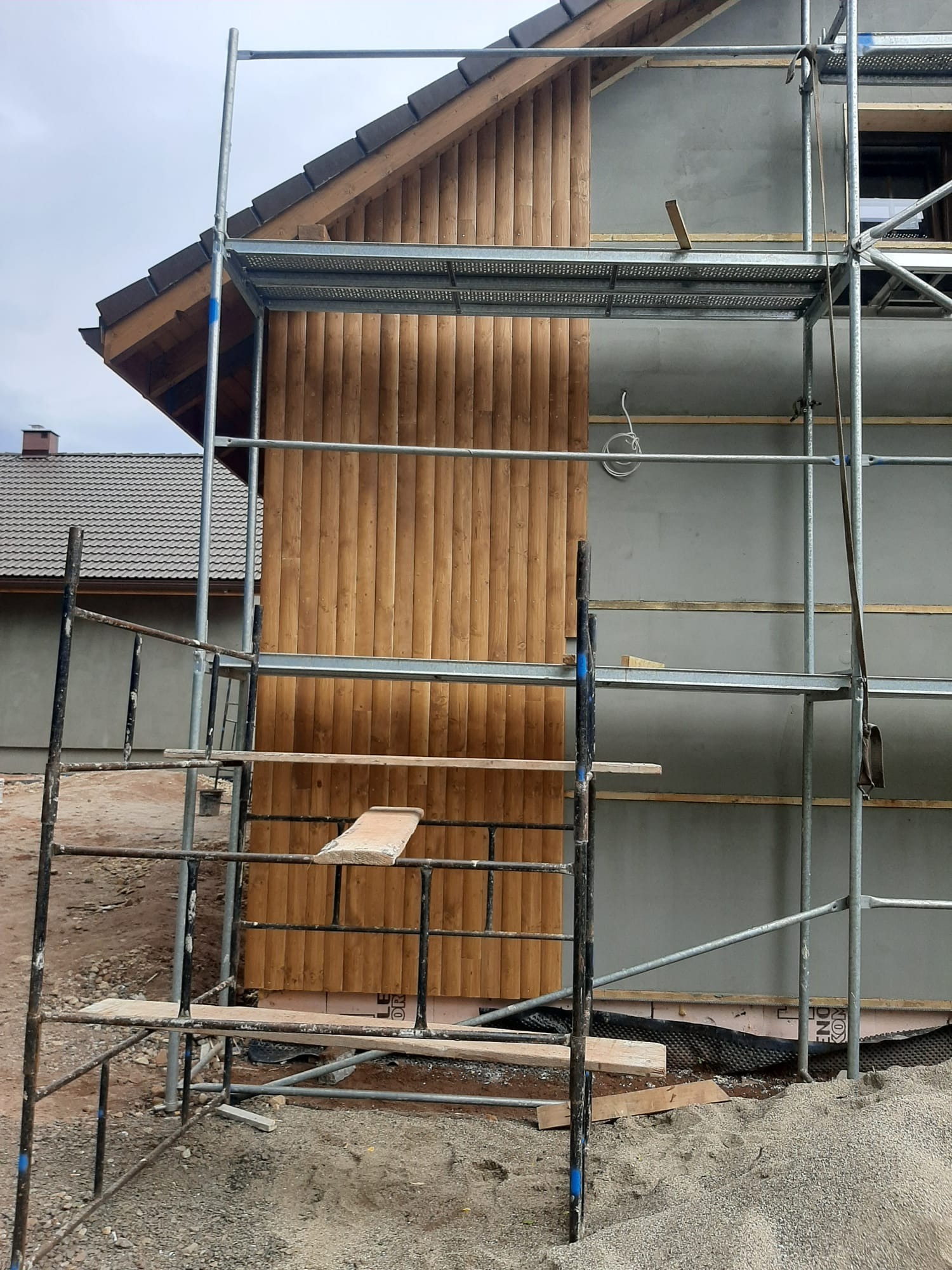 Budowa domu szkieletowego: fragment elewacji z drewnianą boazerią i szarym tynkiem, widoczne rusztowania. Etap prac wykończeniowych.