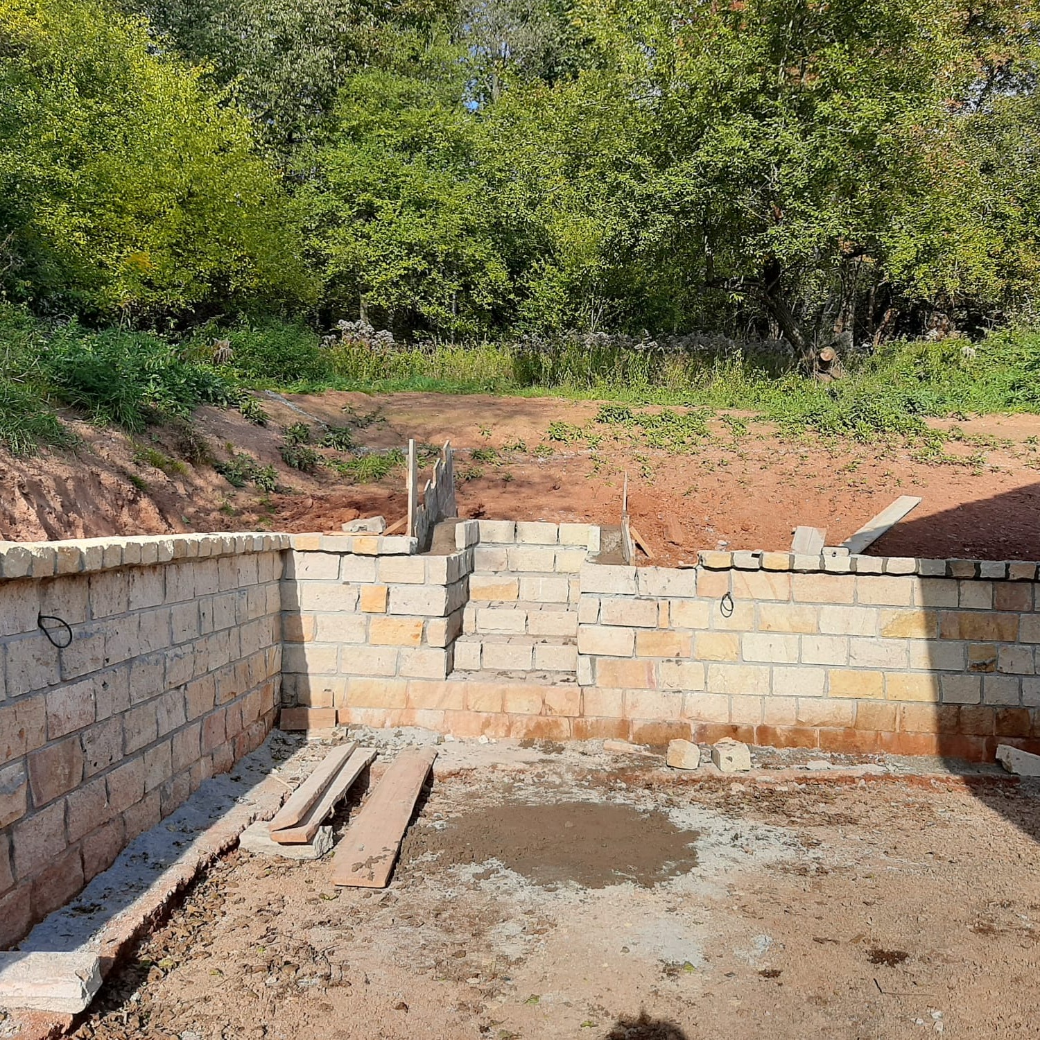 Wznoszenie muru oporowego z jasnych cegieł na nierównym terenie. Widoczne fragmenty konstrukcji i narzędzia. Tło stanowi zieleń drzew i ziemia.