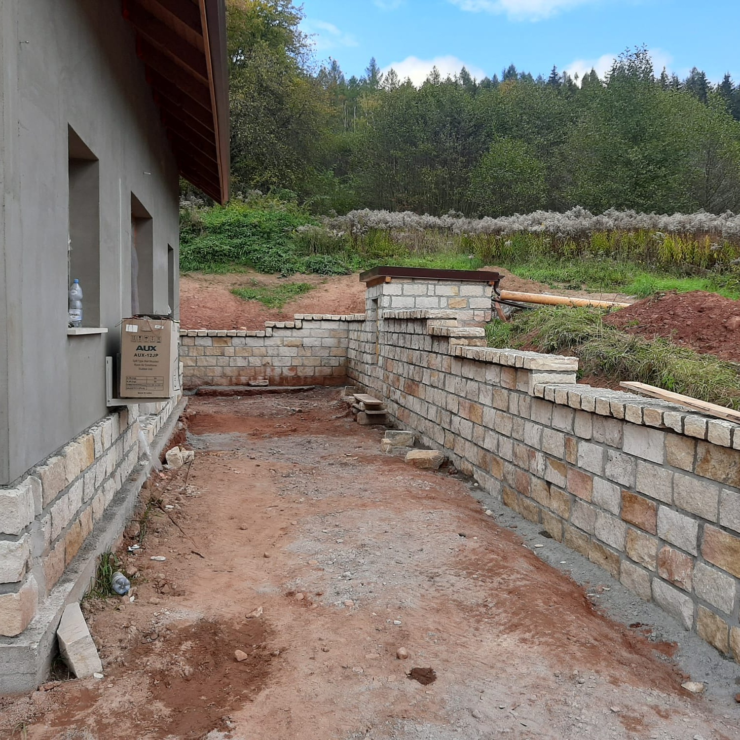 Budynek w trakcie wykończenia z kamienną elewacją i murem oporowym z kamienia. Widoczne elementy konstrukcyjne i otaczający krajobraz.