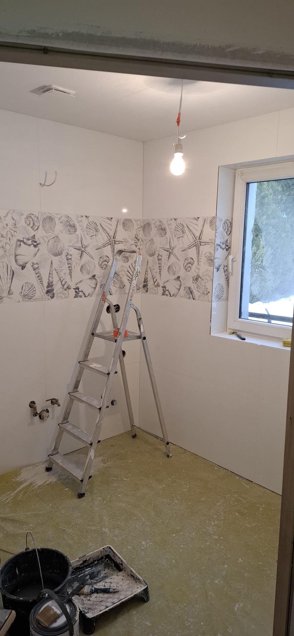 Remont łazienki: białe płytki z motywem morskim, drabina malarska, widoczne rury i narzędzia malarskie na zabezpieczonej podłodze. W tle okno.