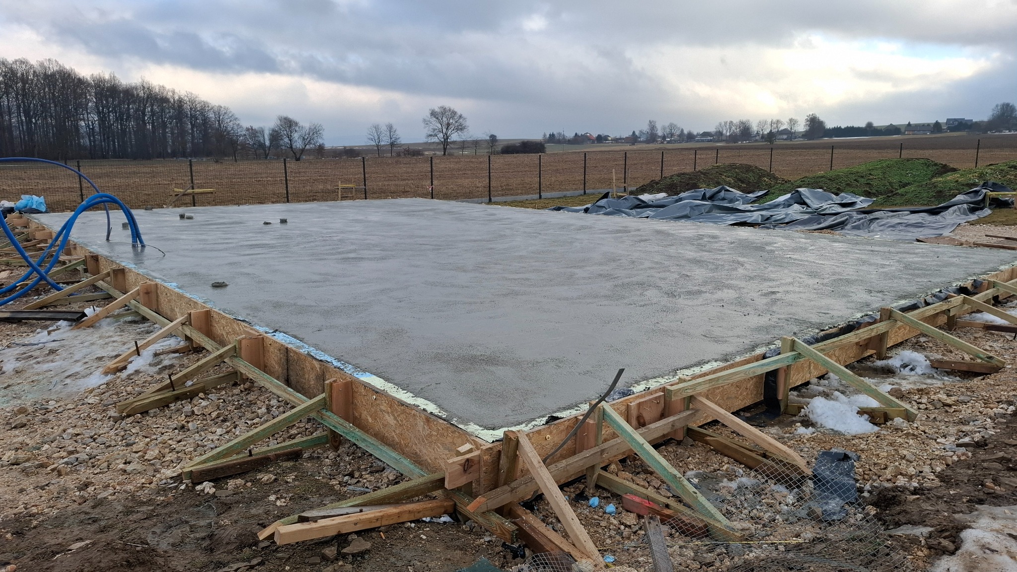 Zalany betonem fundament z drewnianym szalunkiem, widoczne niebieskie rury i otaczający teren z resztkami śniegu. Pochmurne niebo w tle.