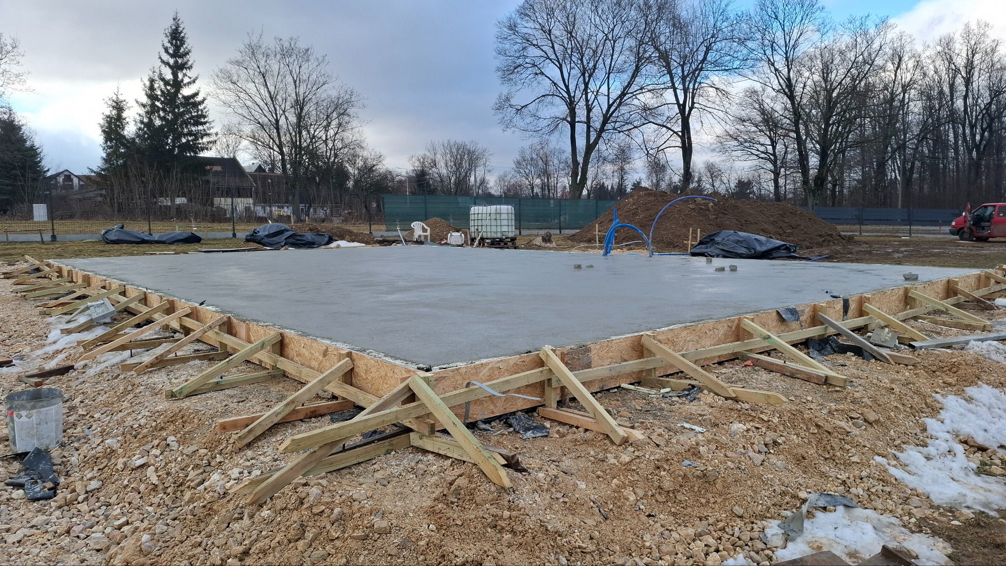 Zalany betonowy fundament z drewnianym szalunkiem, widok z poziomu gruntu. W tle drzewa i częściowo widoczny czerwony samochód.
