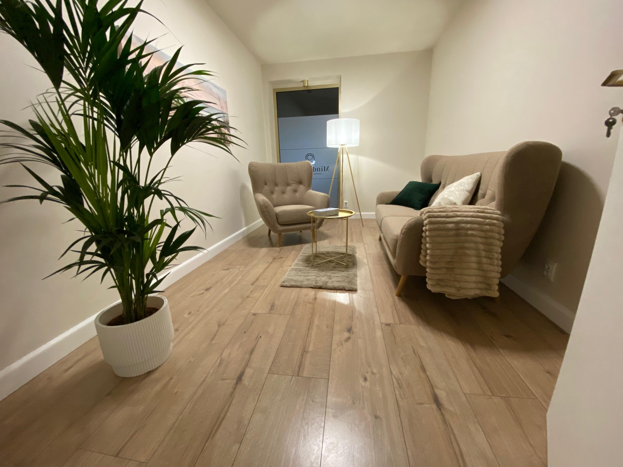 Jasne wnętrze gabinetu z beżowymi meblami, lampą stojącą i rośliną doniczkową. Przytulna aranżacja z drewnianą podłogą i subtelnymi dekoracjami.