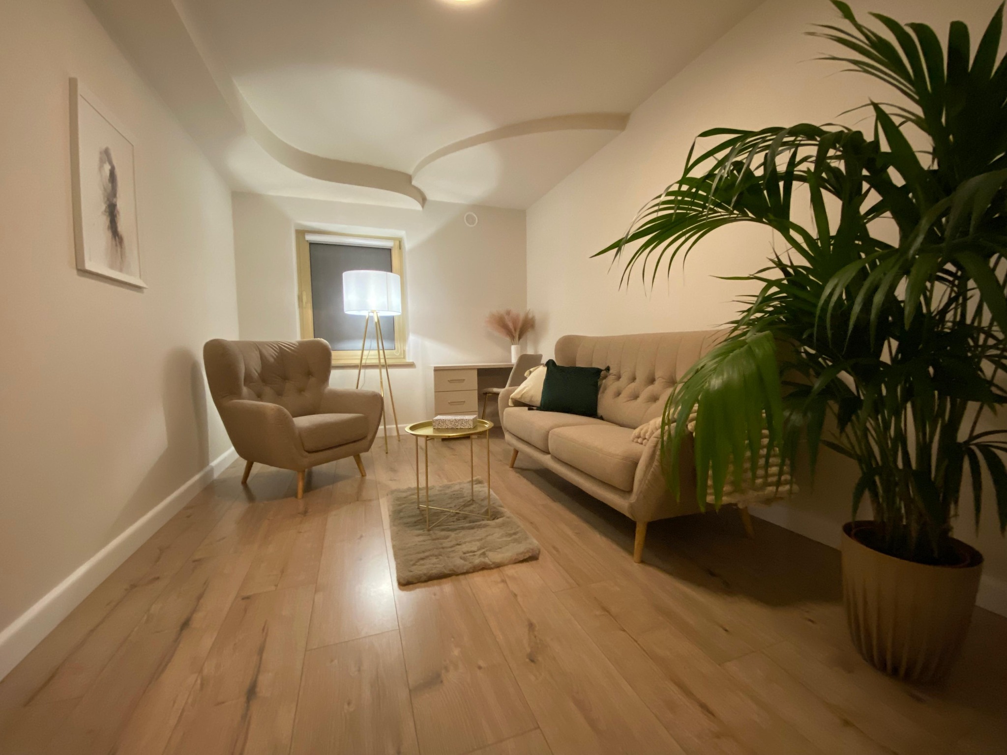 Jasne wnętrze gabinetu z beżowym fotelem, sofą i rośliną doniczkową. Na ścianie obraz, a na suficie dekoracyjne wykończenie. Przytulna aranżacja.