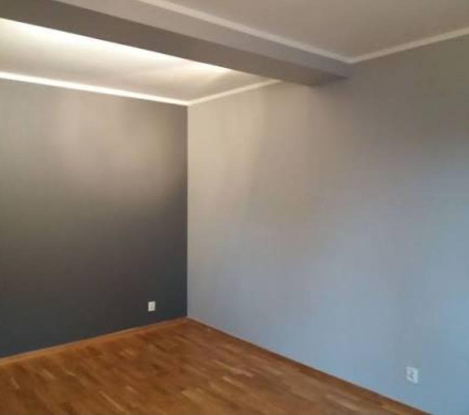 Pokój po remoncie: parkiet, szare ściany z listwami, widoczne gniazdka. Minimalistyczne wnętrze po odświeżeniu, z akcentem na drewnianą podłogę i stonowaną kolorystykę ścian.