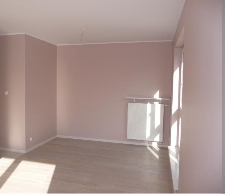 Pusty pokój po remoncie: ściany w odcieniach różu, jasna podłoga, biały grzejnik pod oknem z promieniami słońca. Minimalistyczne wykończenie wnętrza.