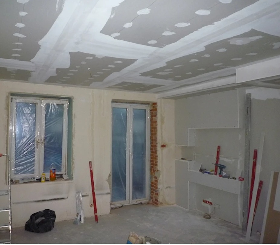 Remontowane wnętrze: sufit z płyt gipsowych z widocznymi łączeniami, ściany w trakcie szpachlowania, okna zabezpieczone folią, narzędzia pracy.