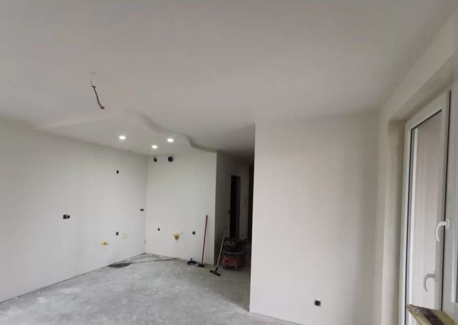 Białe ściany i sufit z oświetleniem punktowym w świeżo wykończonym wnętrzu. Widoczne gniazdka elektryczne i sprzęt budowlany. Okno z białymi ramami po prawej.