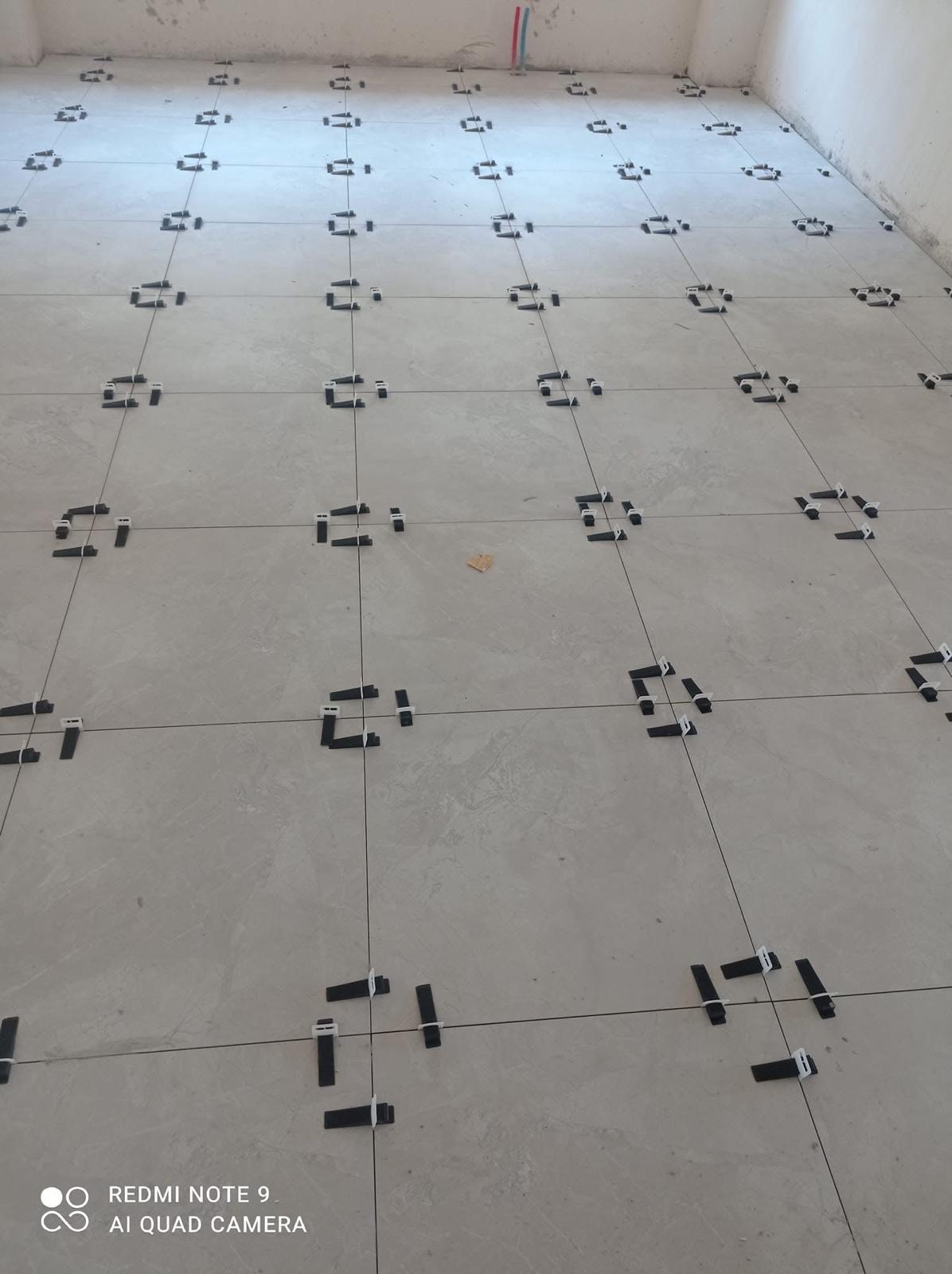 Jasne płytki podłogowe z systemem poziomowania, widoczne fugi i krzyżyki dystansowe. Ujęcie z góry, perspektywa zbieżna. W tle fragment ściany.