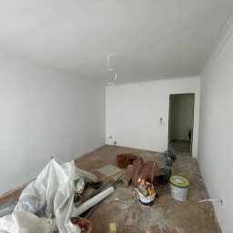 Dawid Piecha - Białe ściany po malowaniu w pustym pokoju z listwami dekoracyjnymi pod sufitem. Na podłodze widoczne narzędzia i resztki materiałów budowlanych.
