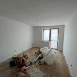 Dawid Piecha - Białe ściany po malowaniu w pokoju z zabezpieczonym oknem. Na podłodze widoczne narzędzia i akcesoria malarskie, co sugeruje etap prac wykończeniowych.