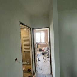 Dawid Piecha - Wnętrze w trakcie remontu: widok na dwa pomieszczenia przez surowe otwory drzwiowe. Ściany po szpachlowaniu, widoczne przewody elektryczne i narzędzia. Podłoga częściowo wykończona.