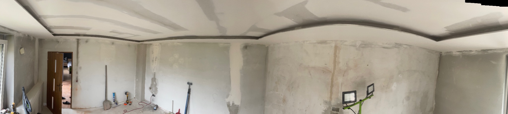 Pokój w trakcie szpachlowania z dekoracyjną listwą sufitową. Widoczne narzędzia i lampa oświetlająca ścianę. Szeroki kadr.