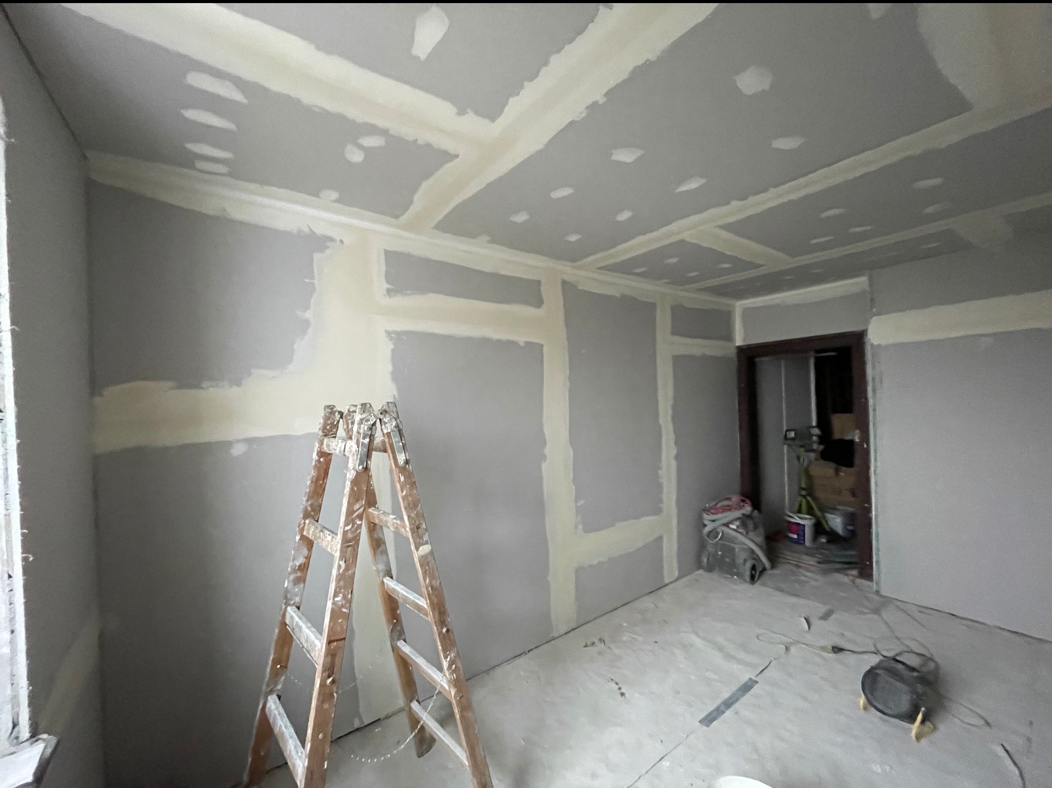 Pokój w trakcie prac wykończeniowych: ściany i sufit pokryte płytami gipsowo-kartonowymi, widoczne łączenia wzmocnione taśmą, drabina malarska i sprzęt budowlany.