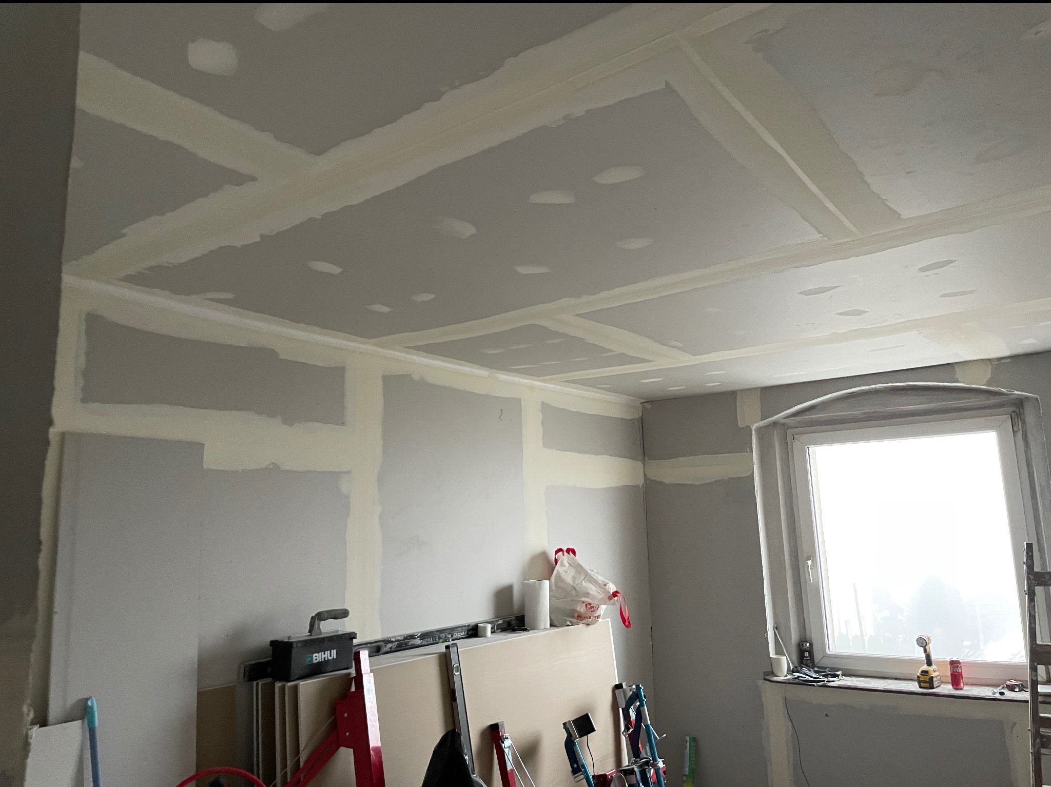 Sufit i ściany po montażu płyt gipsowo-kartonowych i wstępnym szpachlowaniu. Widoczne narzędzia i materiały budowlane. W tle okno z widokiem na zewnątrz.