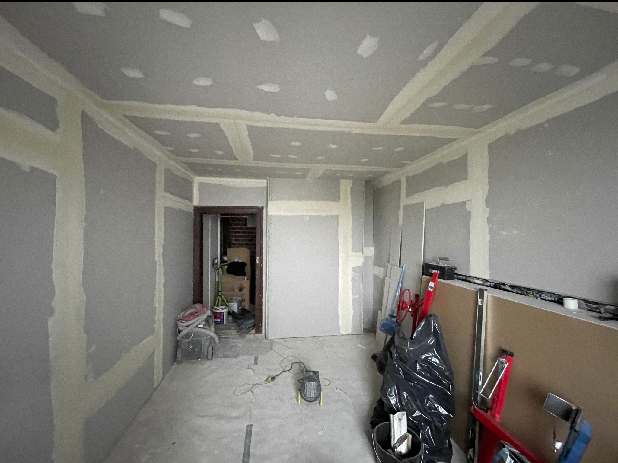 Pokój w trakcie prac wykończeniowych: ściany i sufit pokryte płytami gipsowo-kartonowymi, widoczne łączenia wzmocnione taśmą i szpachlą. Na podłodze narzędzia i materiały budowlane.