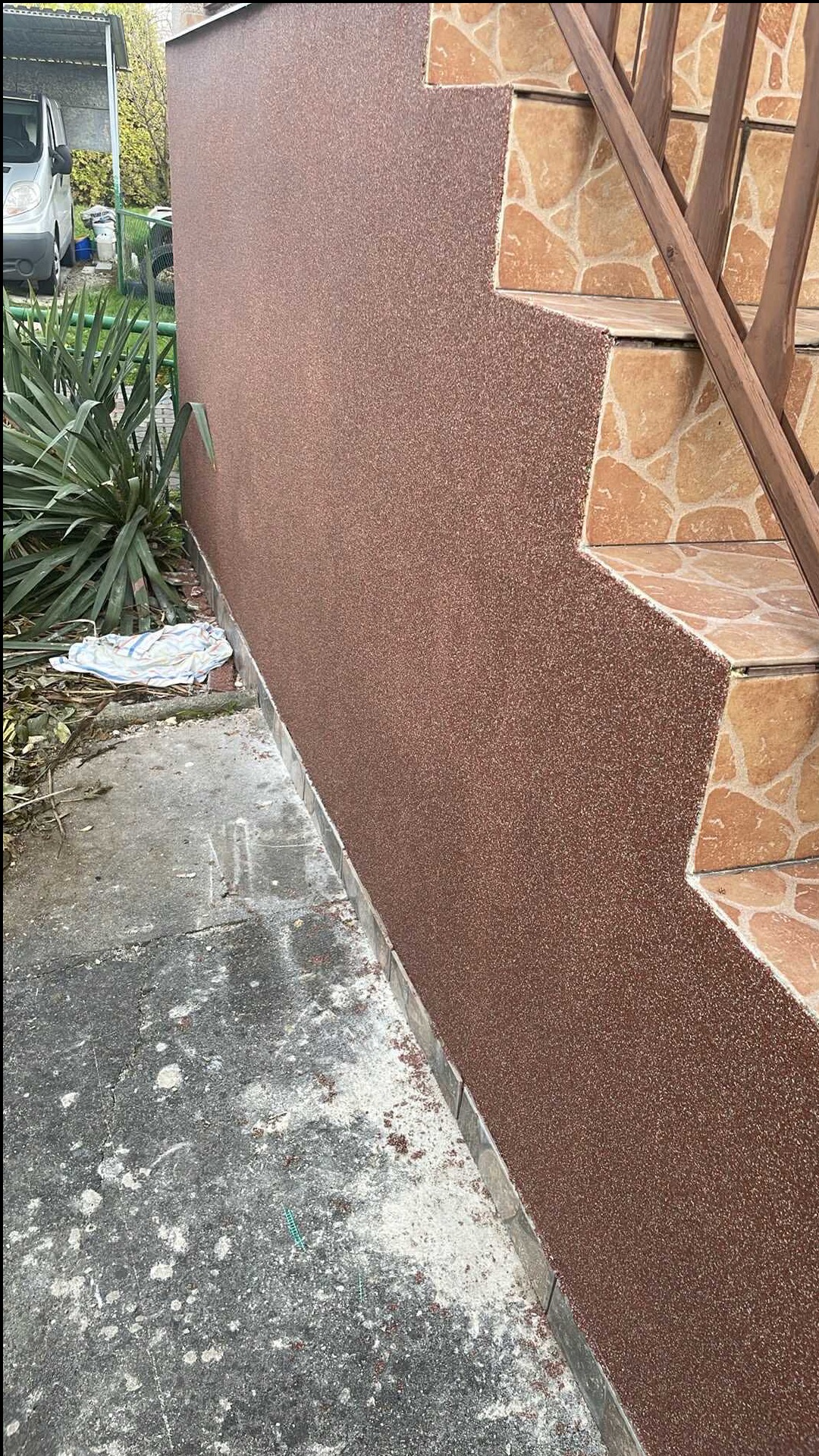 Elewacja budynku z brązowym tynkiem mozaikowym i schodami wyłożonymi płytkami w Rybniku. Widoczne drewniane barierki i fragment chodnika.