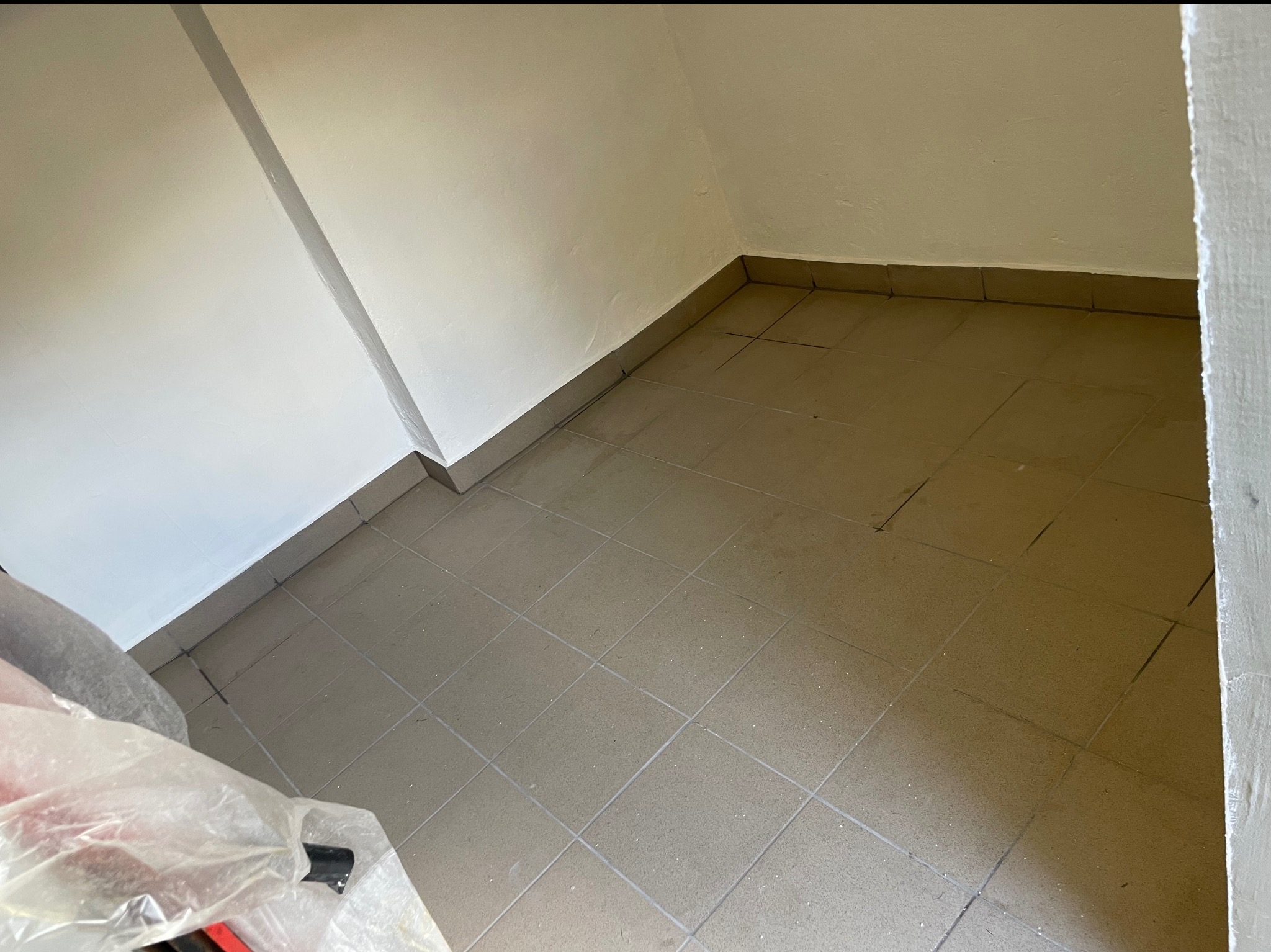 Ukończona podłoga z płytek w rogu pomieszczenia, świeżo po fugowaniu. Widoczne ślady po remoncie na ścianach i podłodze. Ujęcie z góry, blisko podłogi.