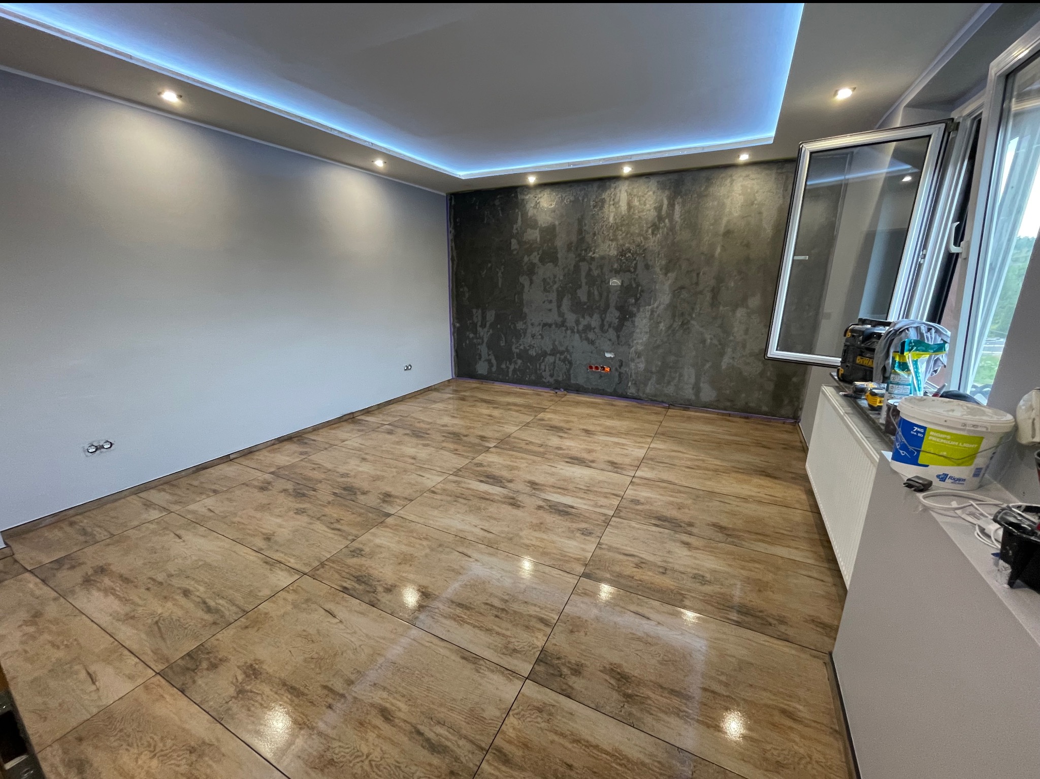 Wykończone wnętrze po remoncie: brązowe płytki podłogowe imitujące drewno, szara ściana i dekoracyjna ściana z efektem betonu. Sufit z oświetleniem LED.