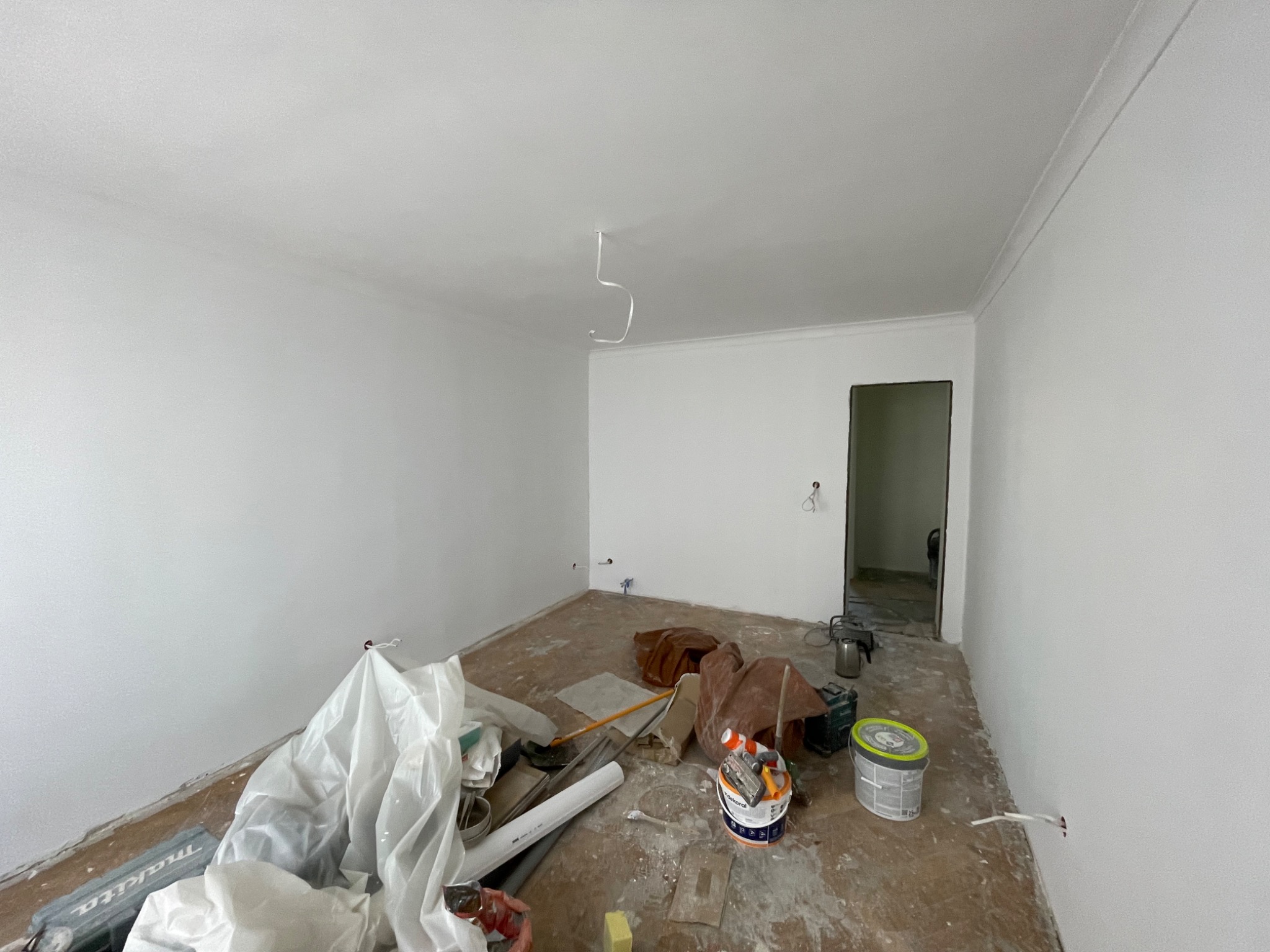 Białe ściany po malowaniu w pustym pokoju z listwami dekoracyjnymi pod sufitem. Na podłodze widoczne narzędzia i resztki materiałów budowlanych.
