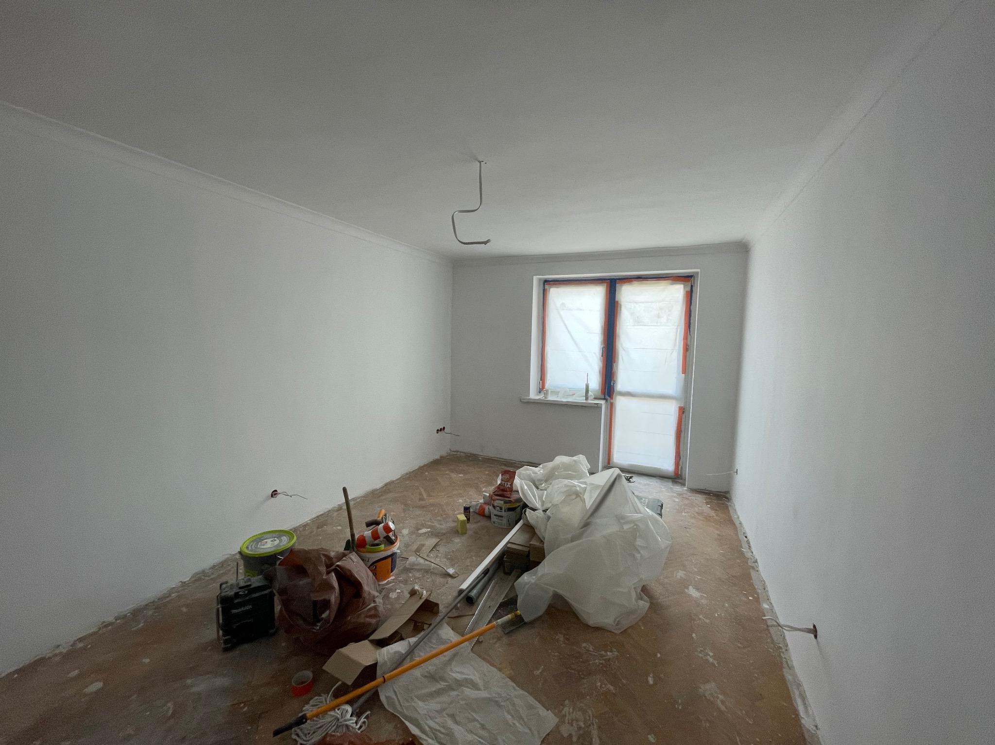 Białe ściany po malowaniu w pokoju z zabezpieczonym oknem. Na podłodze widoczne narzędzia i akcesoria malarskie, co sugeruje etap prac wykończeniowych.