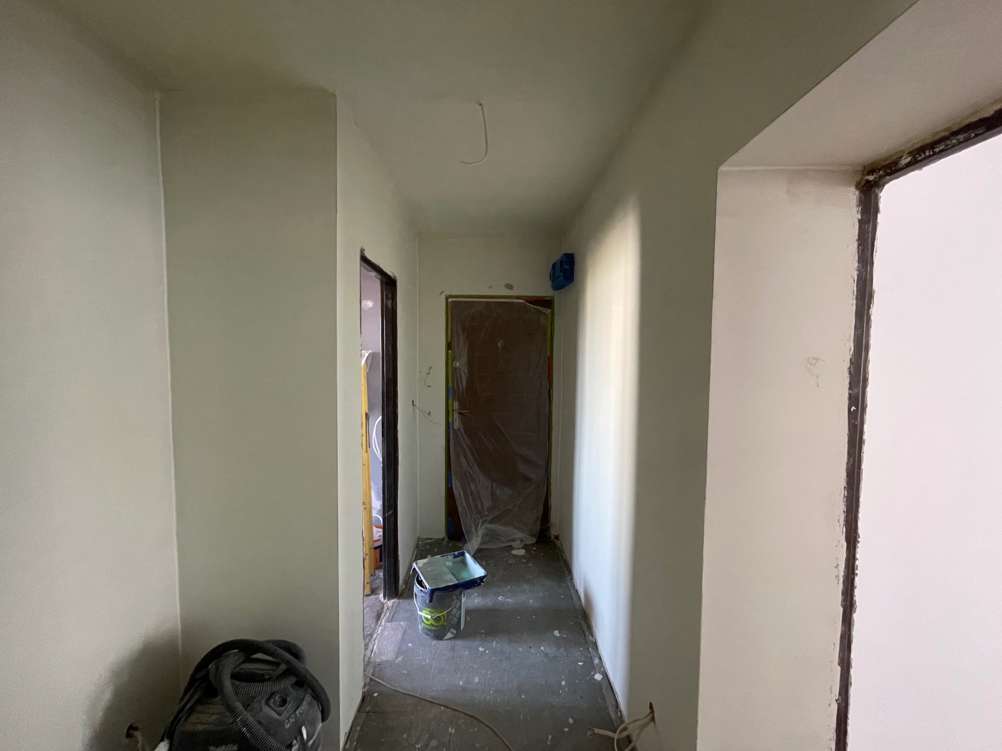 Korytarz po malowaniu na jasny odcień szarości, widoczny sprzęt malarski i zabezpieczone drzwi folią. Świeżo pomalowane ściany i sufit.