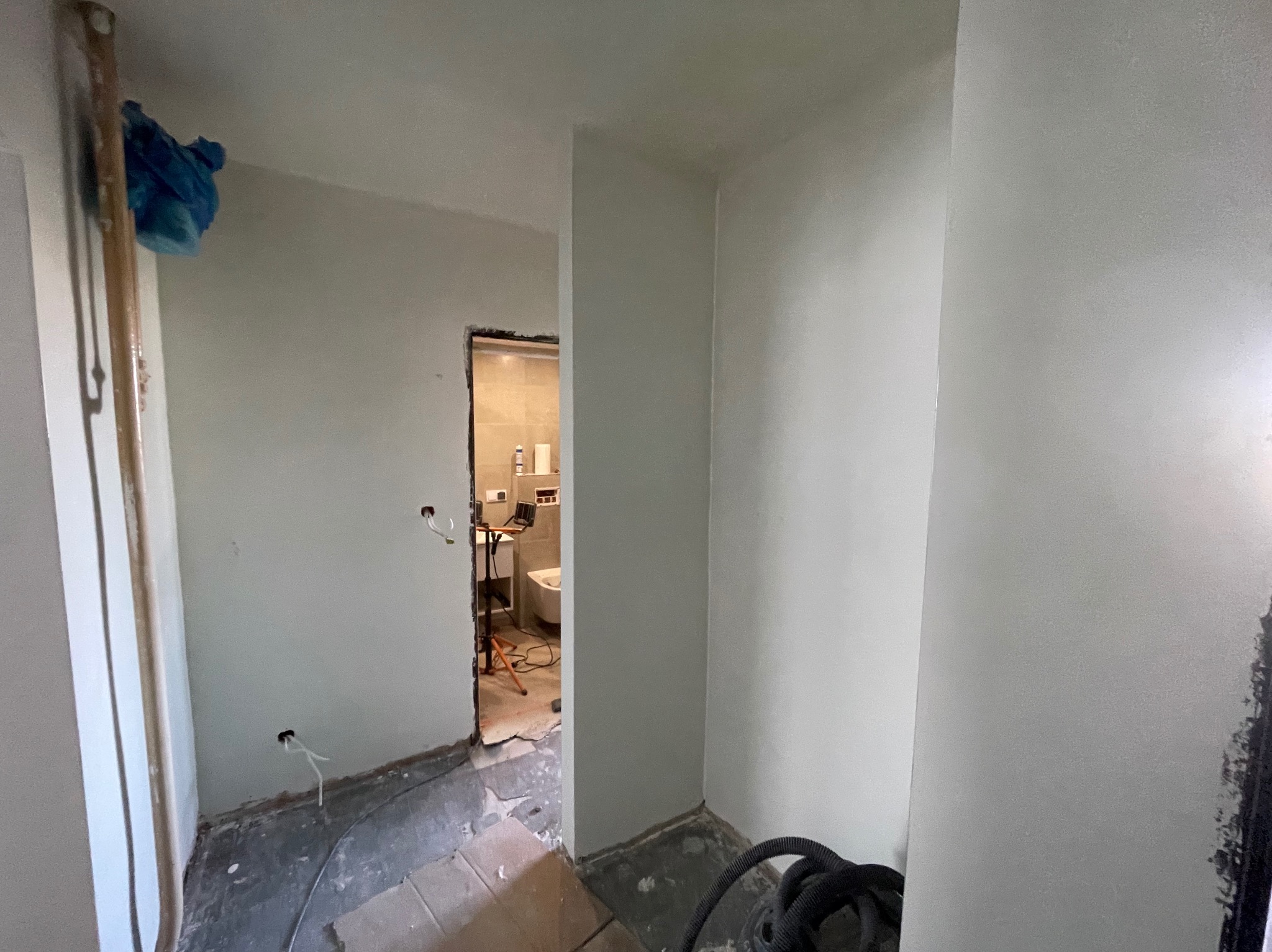 Wnętrze w trakcie remontu: świeżo wyszpachlowane ściany w odcieniach szarości, widoczne kable elektryczne i wejście do wyremontowanej łazienki z białym wyposażeniem.